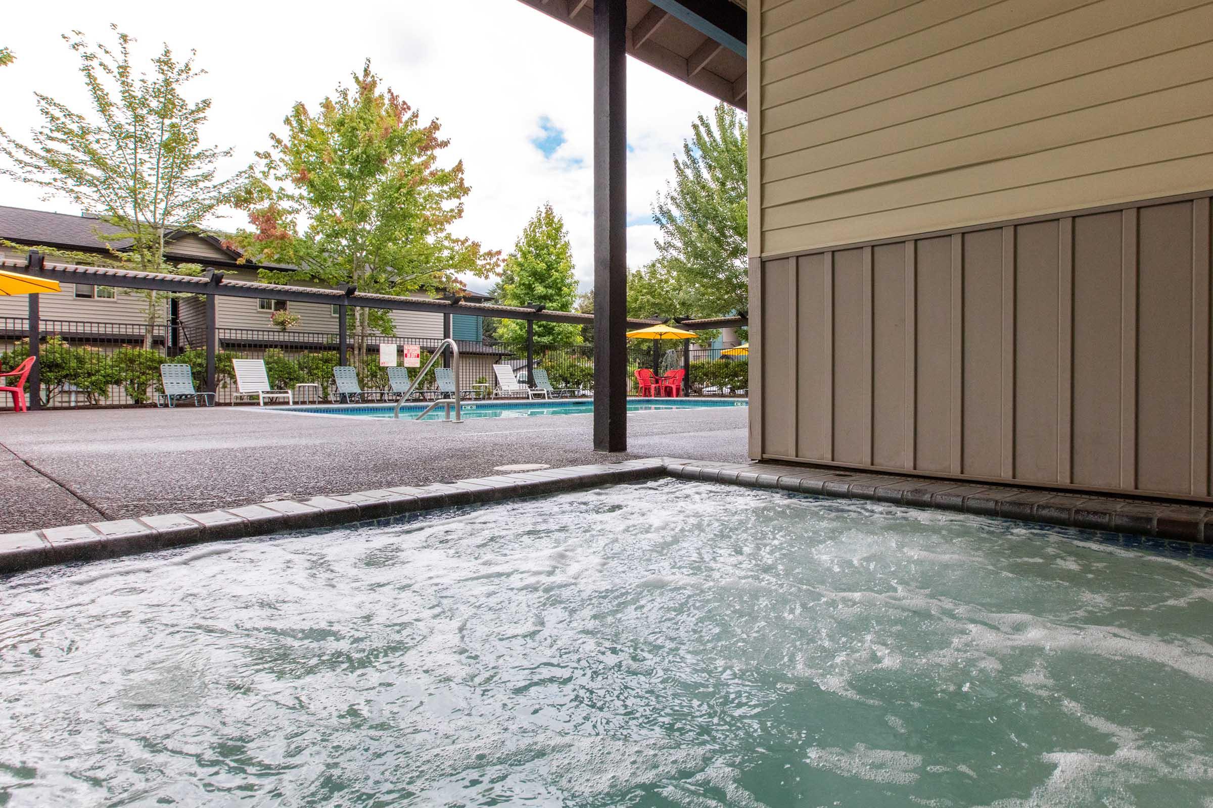 A view from a hot tub overlooking a pool area, featuring green trees and colorful patio umbrellas. The pool is surrounded by lounge chairs, creating a relaxing outdoor atmosphere. A partly cloudy sky adds to the serene setting.
