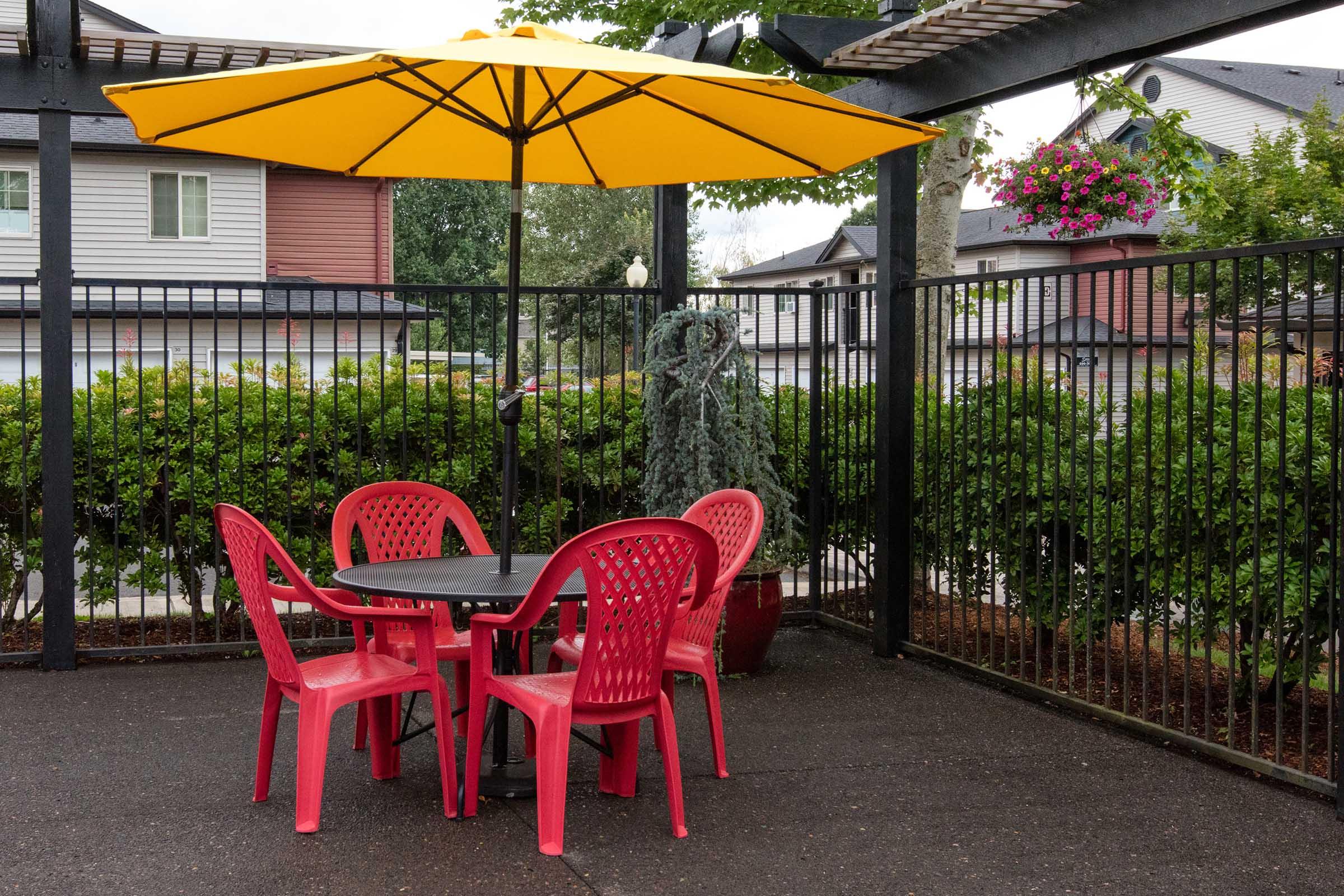 A small outdoor seating area featuring a round black table surrounded by four bright red plastic chairs. An oversized yellow umbrella provides shade, while a green potted plant stands nearby. The backdrop includes a fence and neatly trimmed hedges, with residential buildings visible in the distance under a cloudy sky.