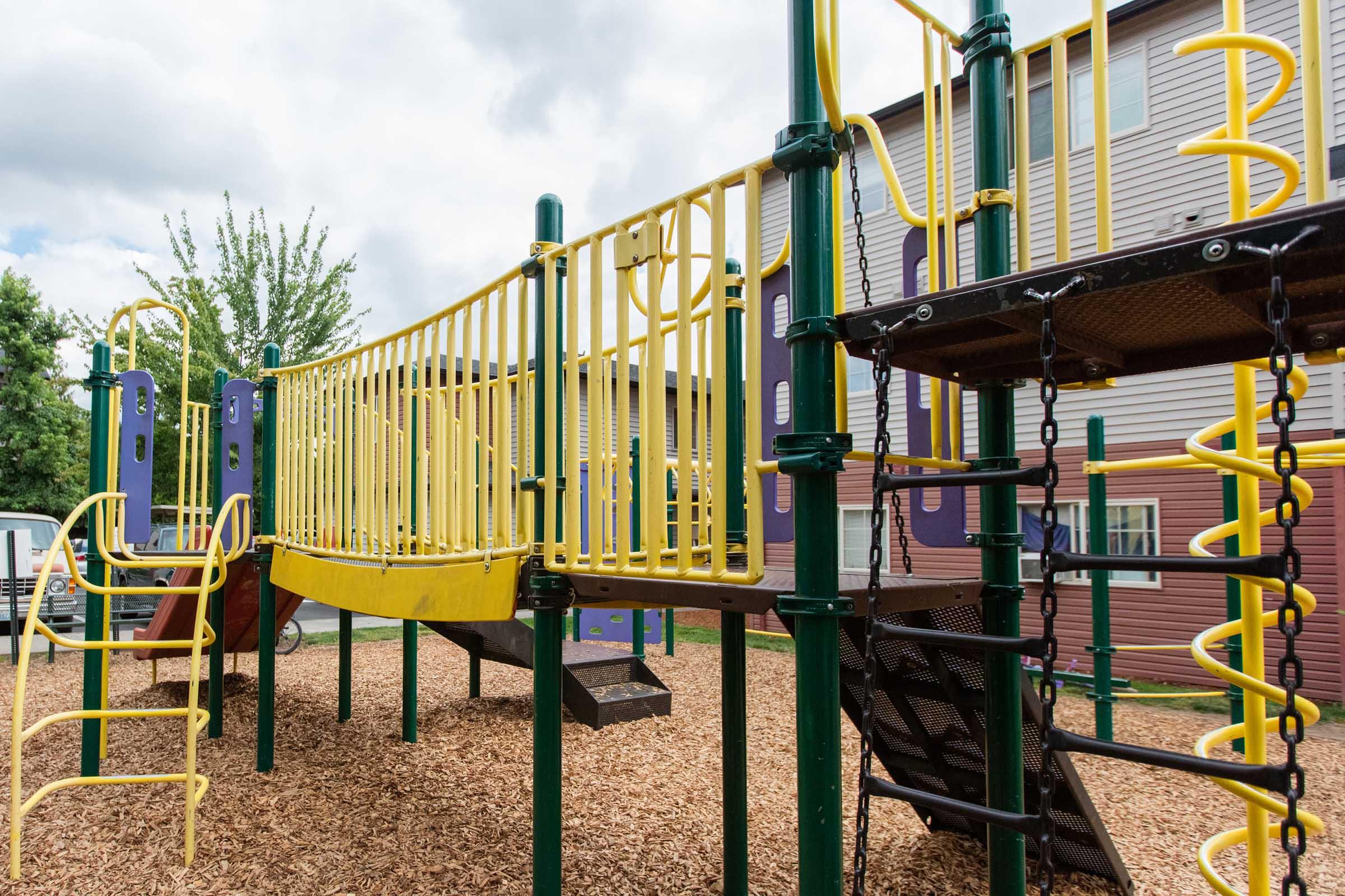 A brightly colored playground featuring slides and climbing structures, surrounded by wood chip flooring. The equipment has yellow railings with climbing frames and a slide, set against a backdrop of trees and a building with multicolored siding. The sky is partly cloudy.