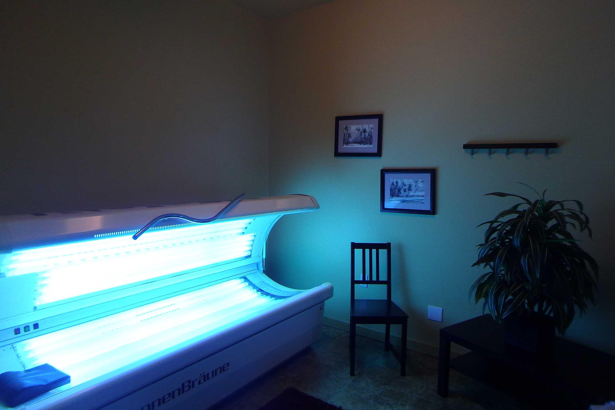 A bright tanning bed in a room with light green walls. A black chair sits nearby next to a small table. Two framed pictures are mounted on the wall, and a potted plant is positioned in the corner. The lighting gives a calm atmosphere, contrasting with the vibrant blue glow of the tanning bed.