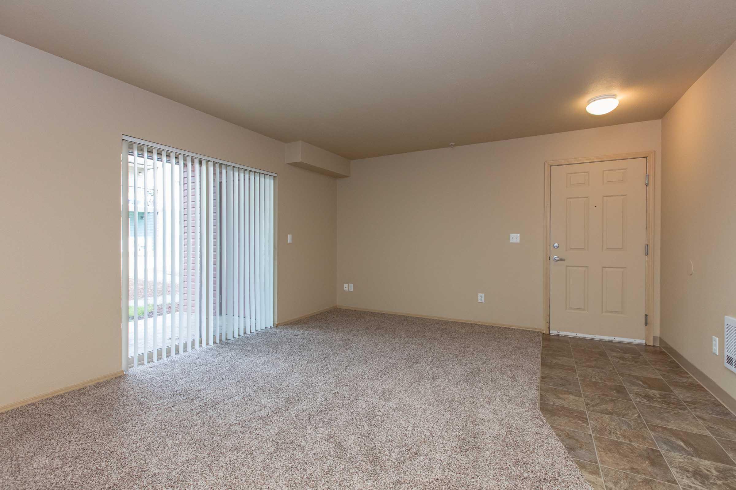 A spacious, empty room with beige walls and carpet flooring. A large window with vertical blinds lets in natural light. The room features a door that leads outside, and light-colored tiled flooring near the entrance. The overall atmosphere is neutral and inviting, suitable for various furnishings.