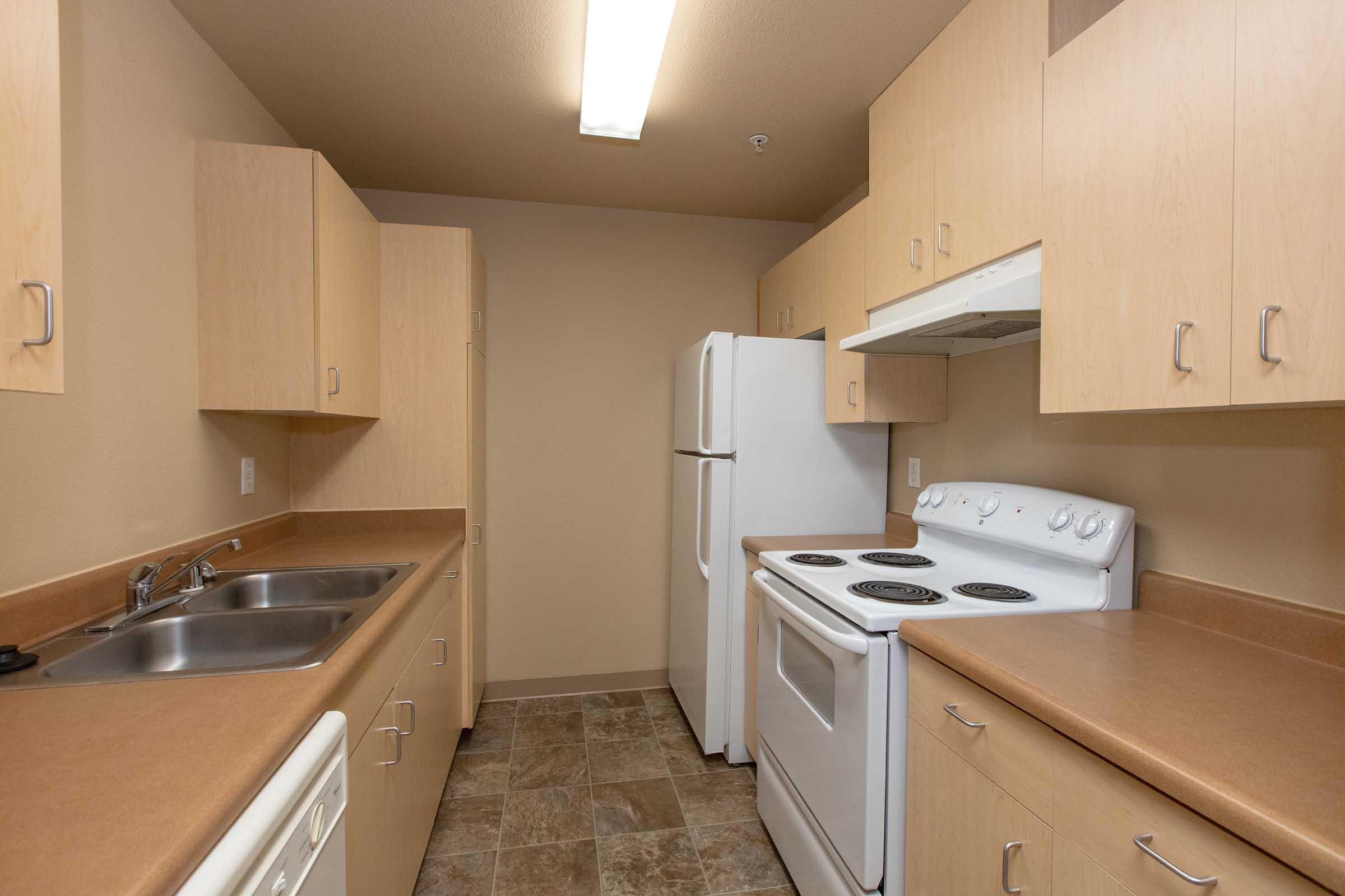 A compact kitchen featuring light wood cabinetry, a double sink, a white refrigerator, an electric stove with oven, and a microwave. The countertop is a warm, solid color, and the floor is tiled in neutral shades. The walls are painted in a soft cream color, creating a bright and inviting atmosphere.