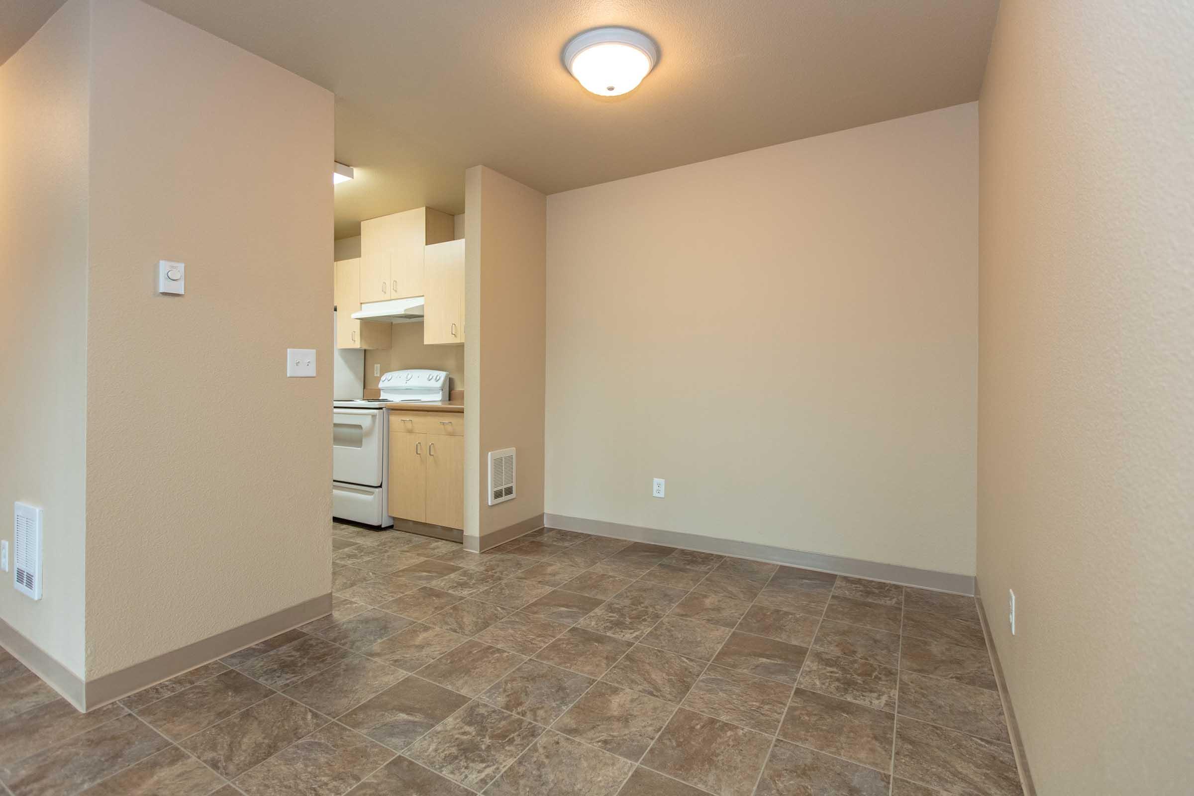 An empty room with light beige walls and a tiled floor. In the back, there is a small kitchen area featuring white cabinetry, a white stove, and an exhaust vent. The ceiling has a round light fixture, and the space is well-lit, creating a spacious and inviting atmosphere.