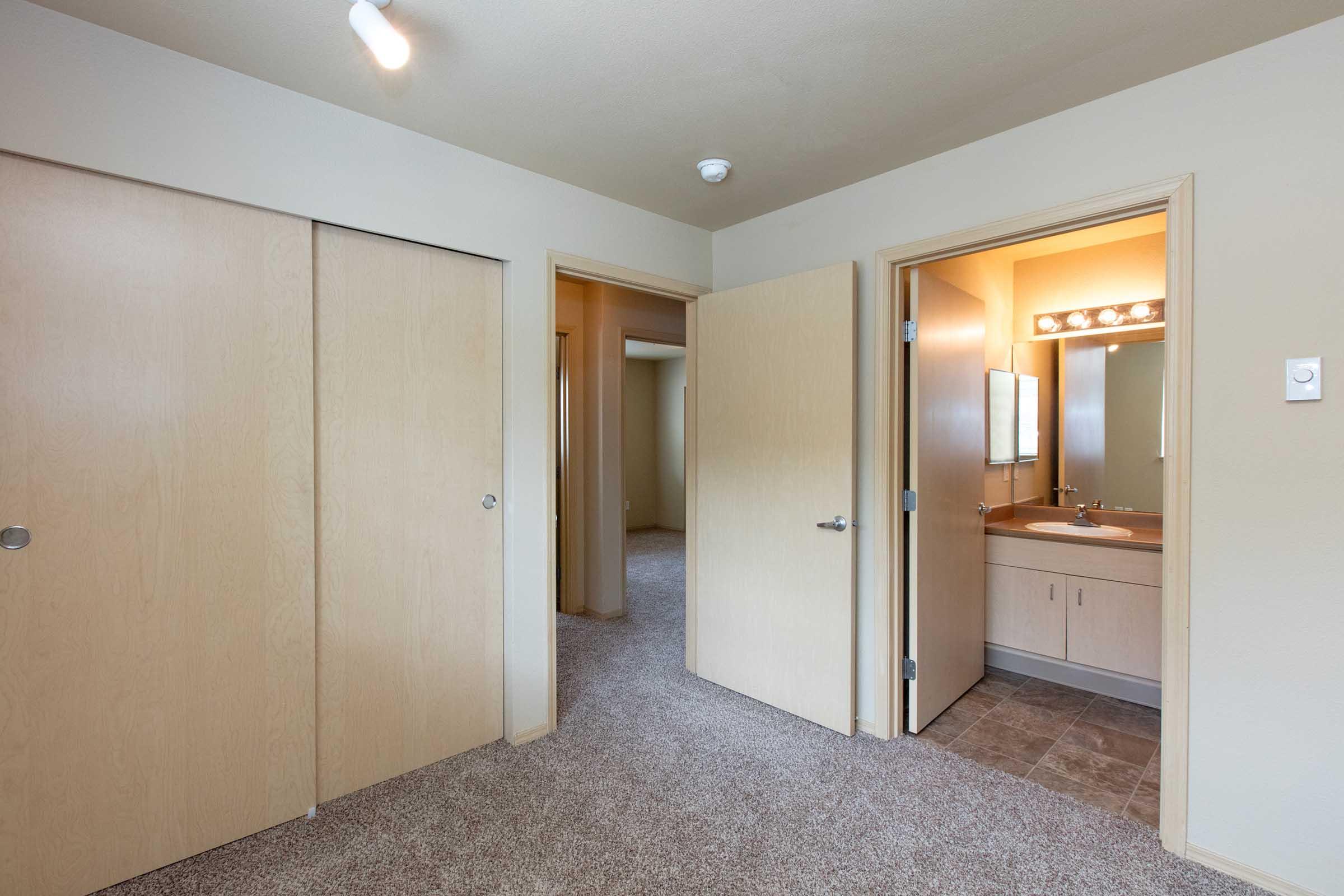 A simple interior view of an apartment hallway featuring beige walls and carpet. To the left, there are two closed closet doors. In the center, an open door leads to a bathroom, which includes a sink and illuminated mirror. Another open doorway in the background suggests access to an additional room.
