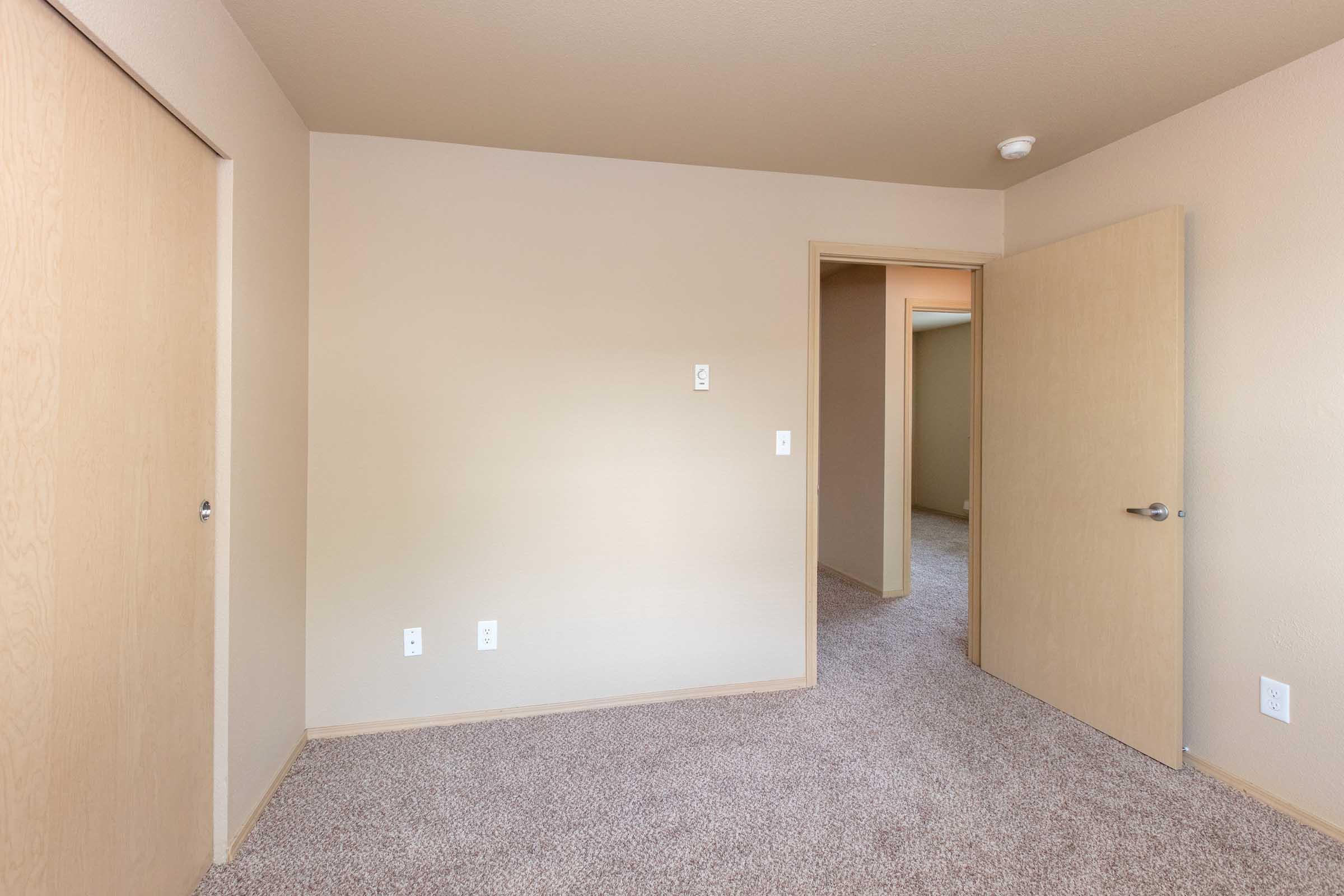 A light beige room with textured carpet, featuring two wooden doors on the left and one door leading to another room in the background. The walls are painted a soft beige, and there is a light fixture on the ceiling. The overall atmosphere is neutral and minimalist.