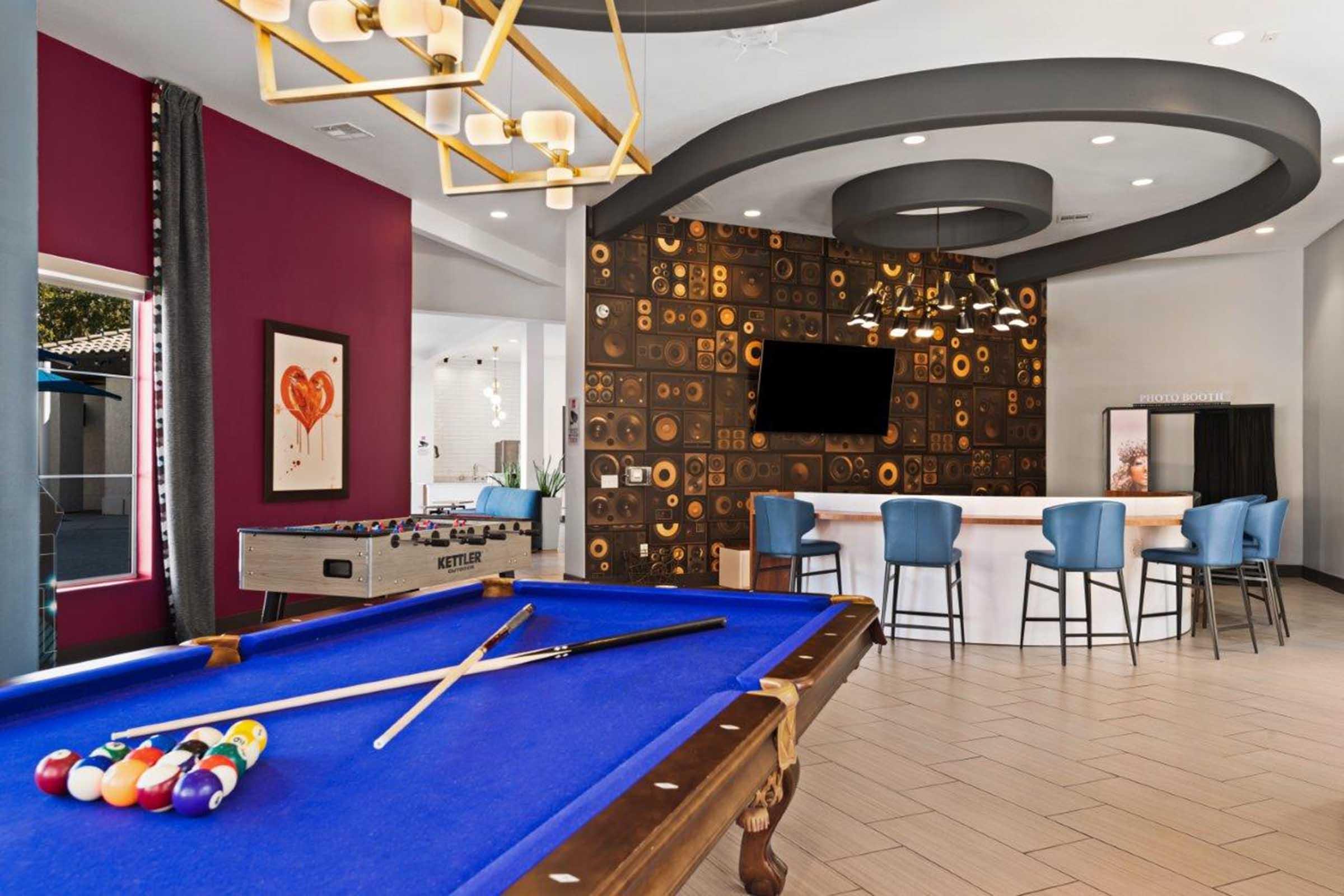 A stylish indoor recreational area featuring a blue pool table with colorful balls, a foosball table, and a modern bar with blue stools. The decor includes textured walls, pendant lighting, and artwork, creating a vibrant and inviting atmosphere for socializing and entertainment.