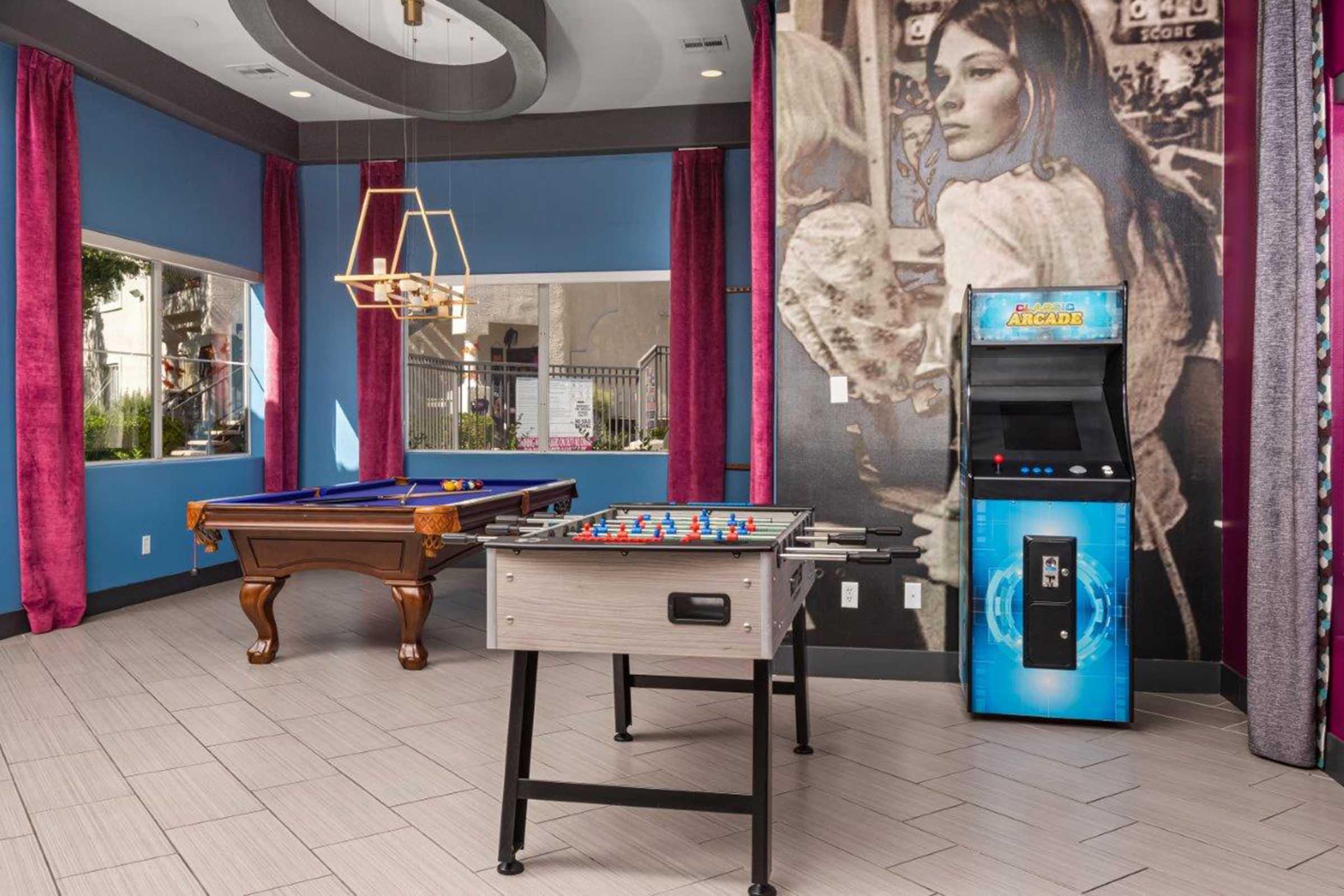 A modern game room featuring a billiard table, a foosball table, and an arcade machine. The walls are adorned with vibrant purple curtains and artwork, creating a lively atmosphere. Large windows allow natural light, enhancing the playful environment for socializing and entertainment.