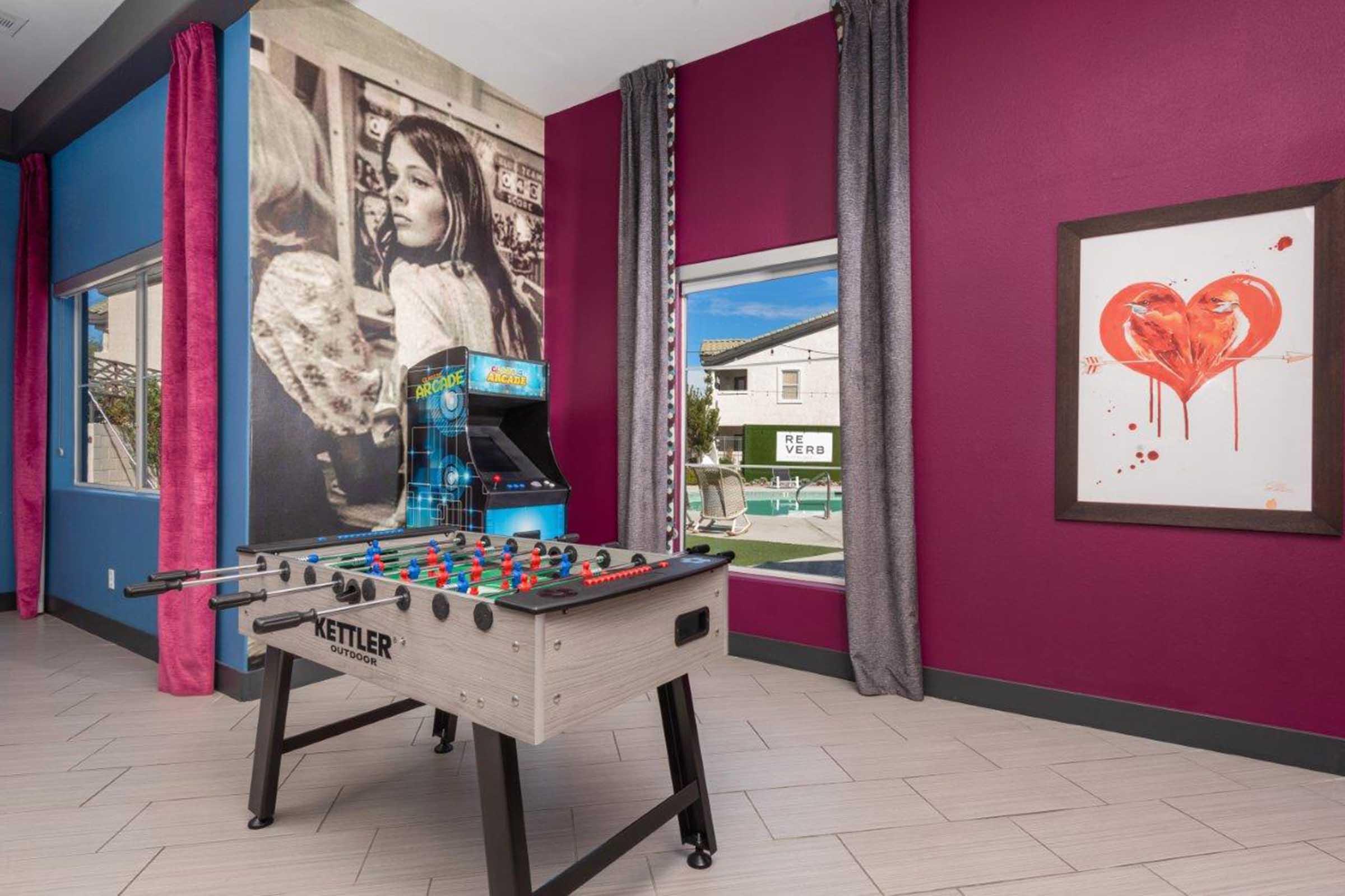 A vibrant game room featuring a foosball table in the foreground, colorful walls in purple and teal, large windows letting in natural light, an arcade game in the background, and wall art depicting a girl and a heart design.