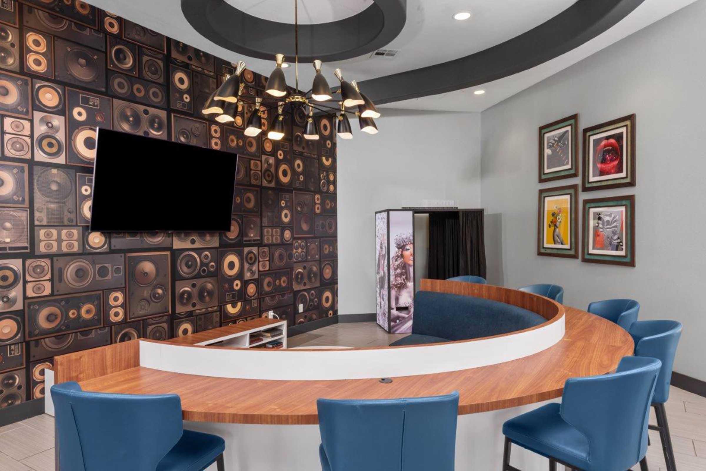 A modern lounge area featuring a curved wooden bar with blue chairs, a wall decorated with speaker patterns, and a large flat-screen TV. Art pieces hang on one wall, and a vending machine is visible in the corner, contributing to a contemporary and stylish atmosphere.