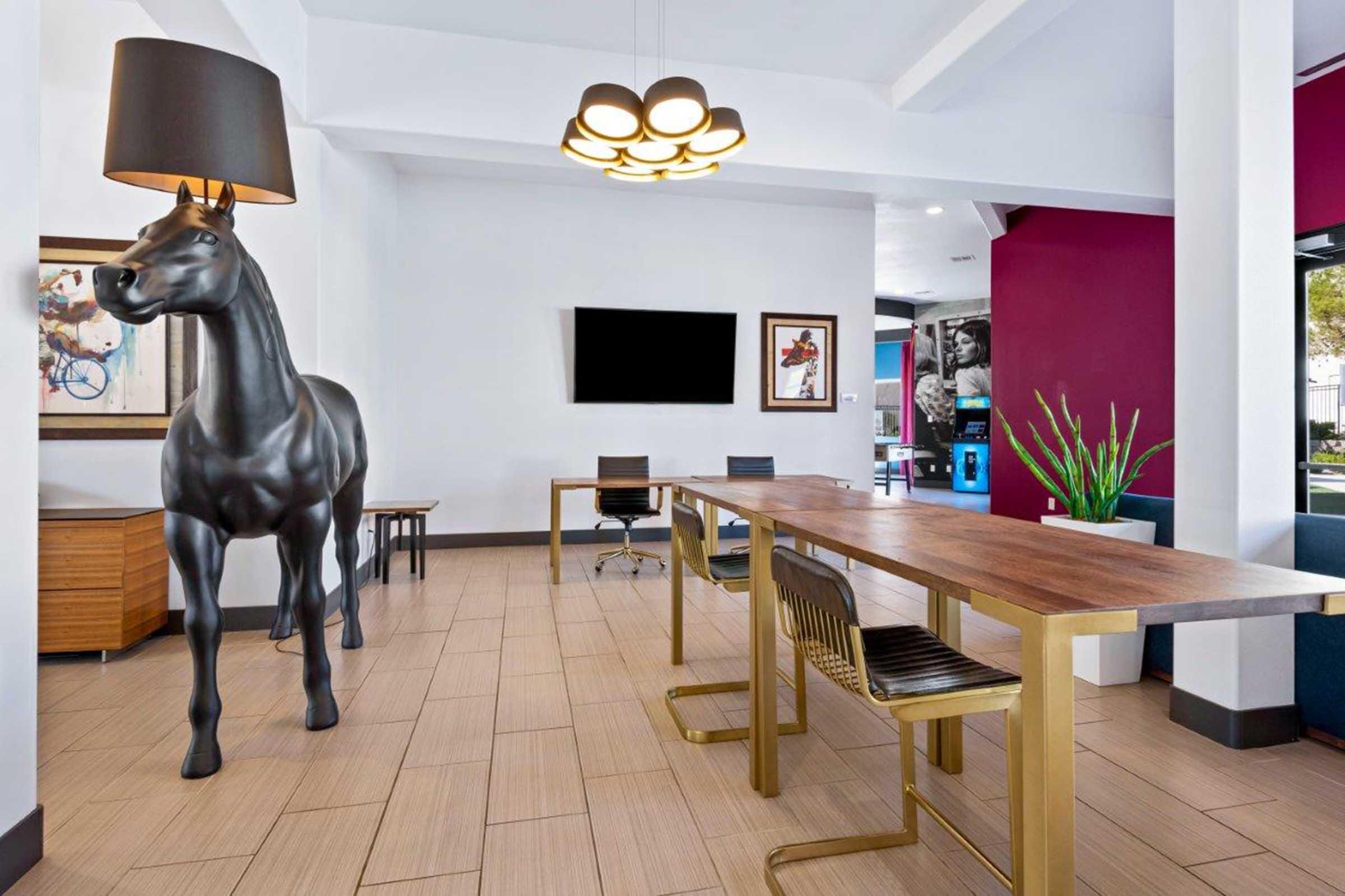 A modern interior space with a large black horse sculpture serving as a lamp. There's a wooden conference table with metallic chairs, and minimalist artwork on the walls. A television is mounted on one wall, and a potted plant adds a touch of greenery. Overall, the decor is stylish and contemporary.