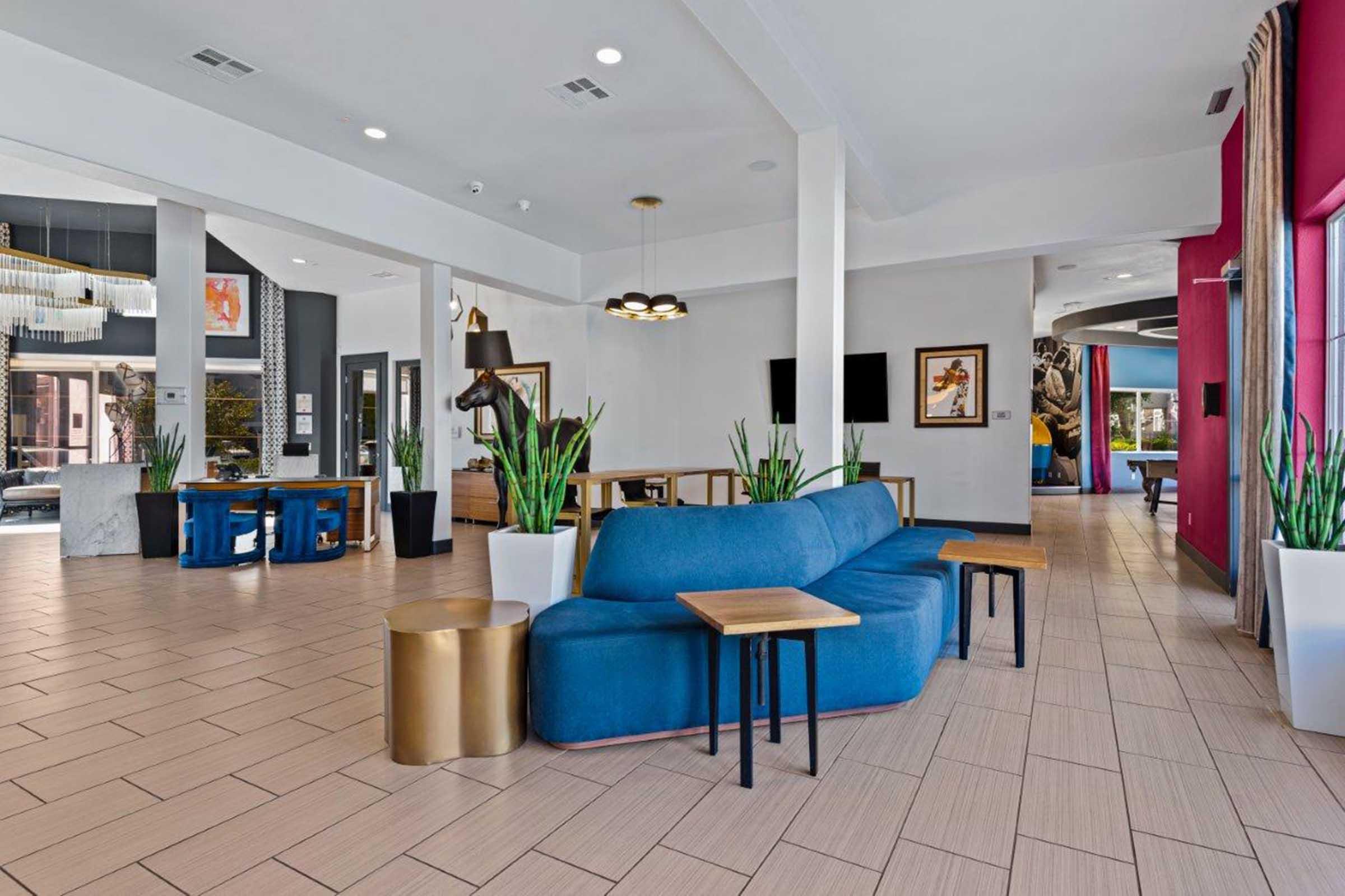 Interior of a modern lobby featuring a blue sectional sofa, wooden side tables, and potted plants. The space has a mix of artwork on the walls and an open layout with natural light. Stylish lighting fixtures and a blend of colors create a welcoming atmosphere.
