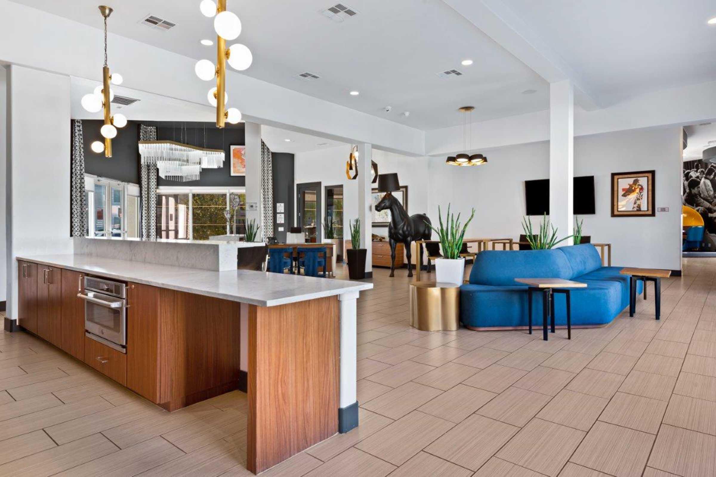 Modern lobby area featuring a long countertop with a kitchen appliance, stylish furniture including a blue couch and round tables. Decorated with art and plants, the space boasts contemporary light fixtures and a large horse statue, creating a welcoming atmosphere.
