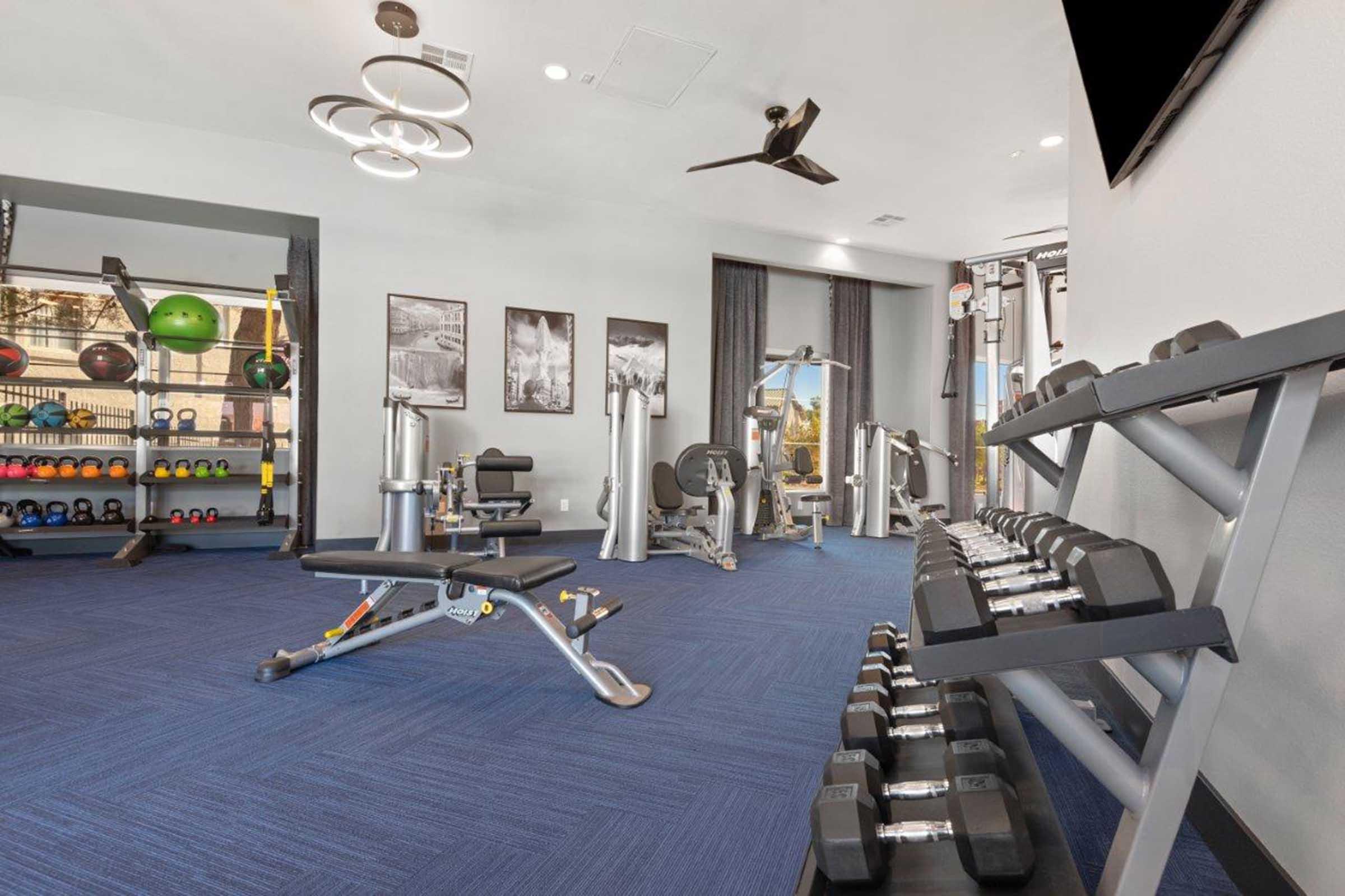 A modern gym interior featuring various fitness equipment, including weight benches, dumbbells on a rack, and workout machines. The space is well-lit with large windows and colorful exercise balls, creating an inviting atmosphere for workouts.