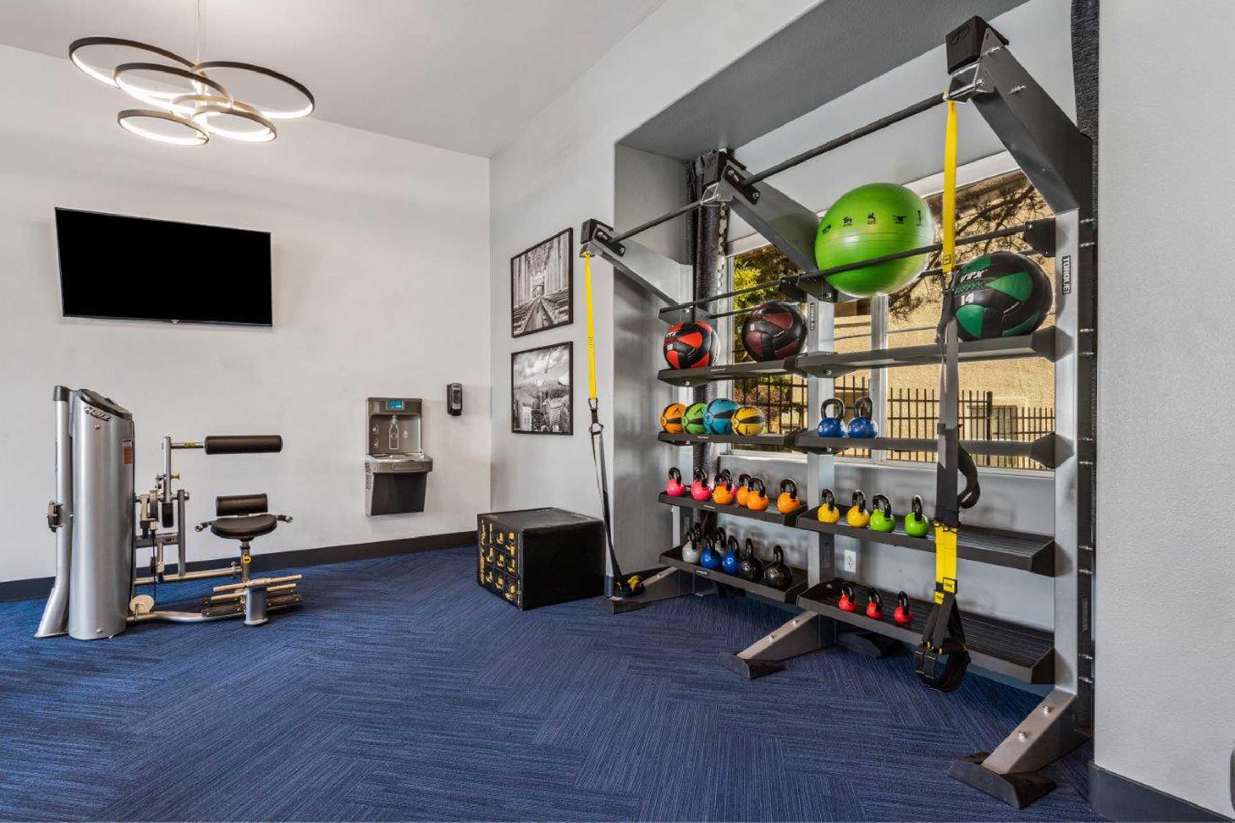 A modern fitness area featuring a variety of exercise equipment. The wall-mounted setup includes colorful kettlebells and a green medicine ball. Nearby, a weight machine is positioned, along with a water cooler and a TV on the wall. A spacious design with blue carpet adds to the gym's ambiance.