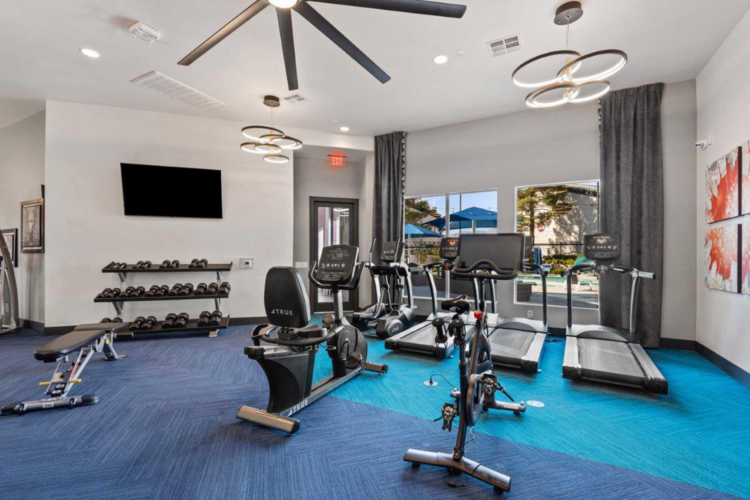 A modern fitness room featuring exercise equipment including treadmills, stationary bikes, and weights. The space has a stylish design with blue carpet, large windows allowing natural light, and contemporary lighting fixtures. A flat-screen TV is mounted on the wall.