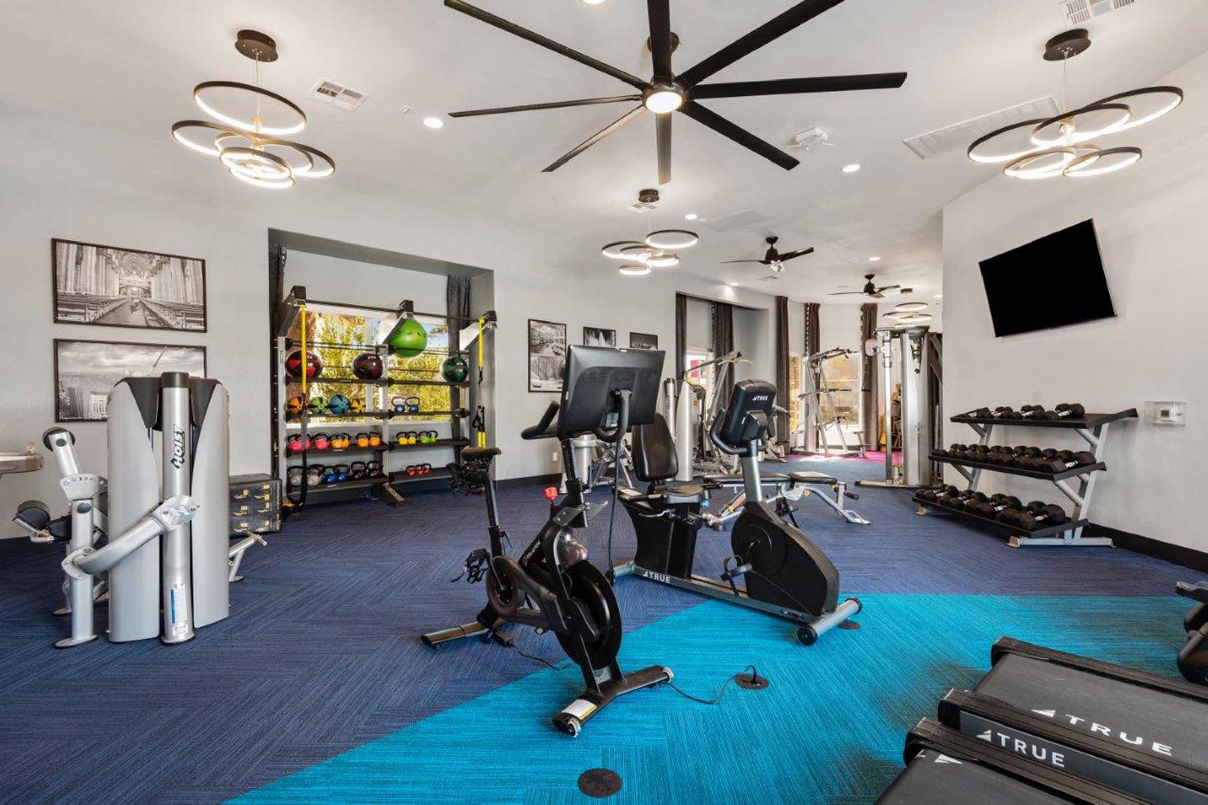 A modern fitness room featuring exercise equipment, including treadmills, elliptical machines, and various weights. The space is well-lit with stylish lighting and has a blue carpet. Wall-mounted screens display fitness information, and there are colorful kettlebells and other workout accessories on display.