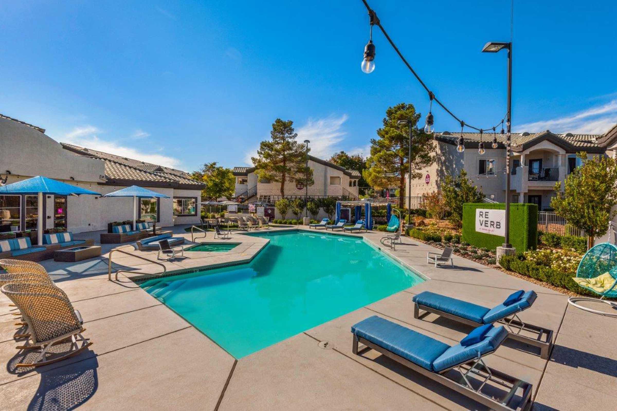 A bright and inviting pool area surrounded by lounge chairs and umbrellas. The pool features a modern design, with landscaped greenery and attractive lighting. Nearby buildings add to the community vibe, creating a relaxing outdoor space perfect for leisure and socializing.