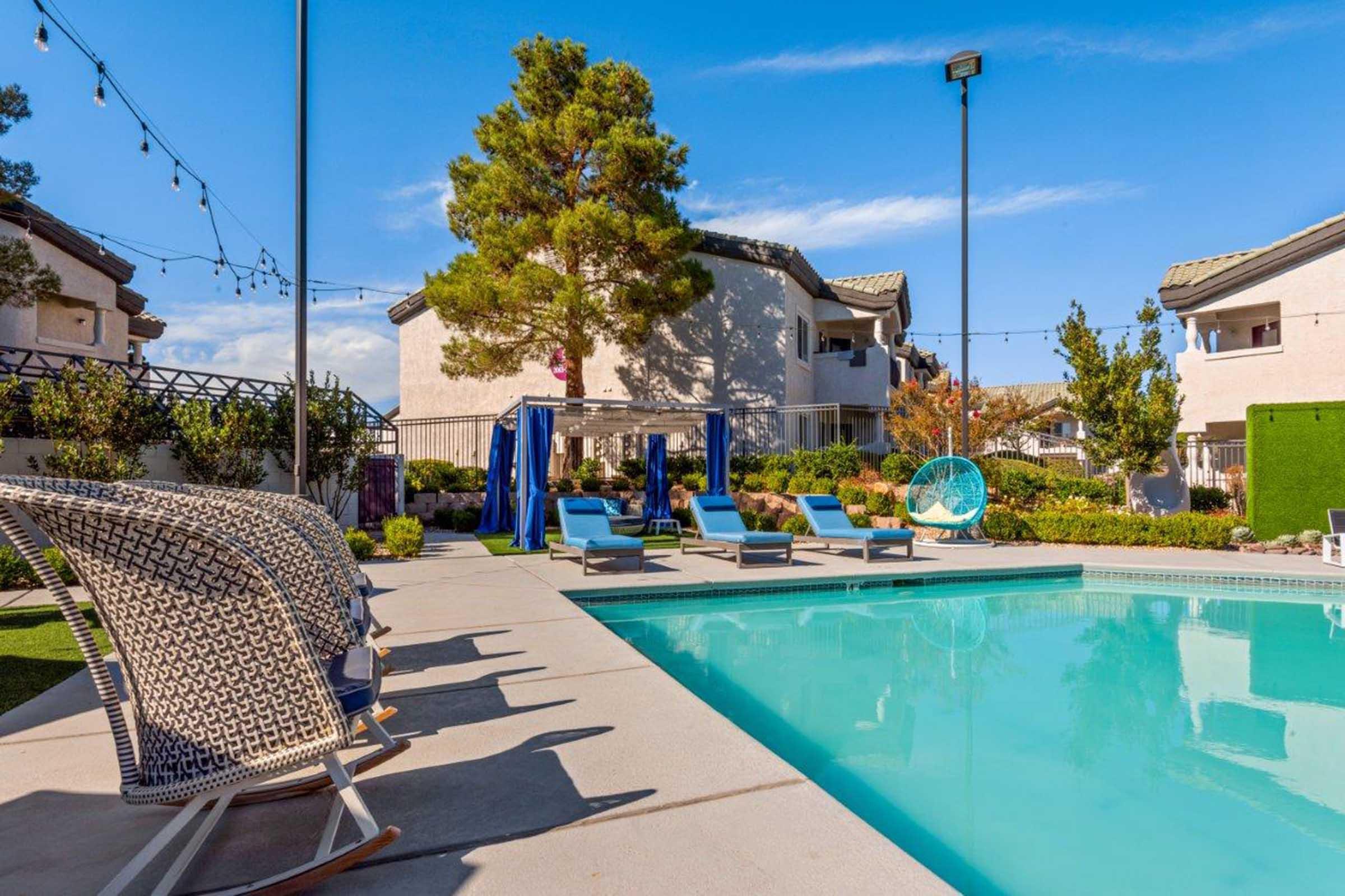 A serene pool area surrounded by lounge chairs and greenery, with blue cabanas in the background. The clear water reflects the blue sky, and string lights are hung above, adding a relaxed and inviting atmosphere. Nearby buildings and trees complete the scenic setting.