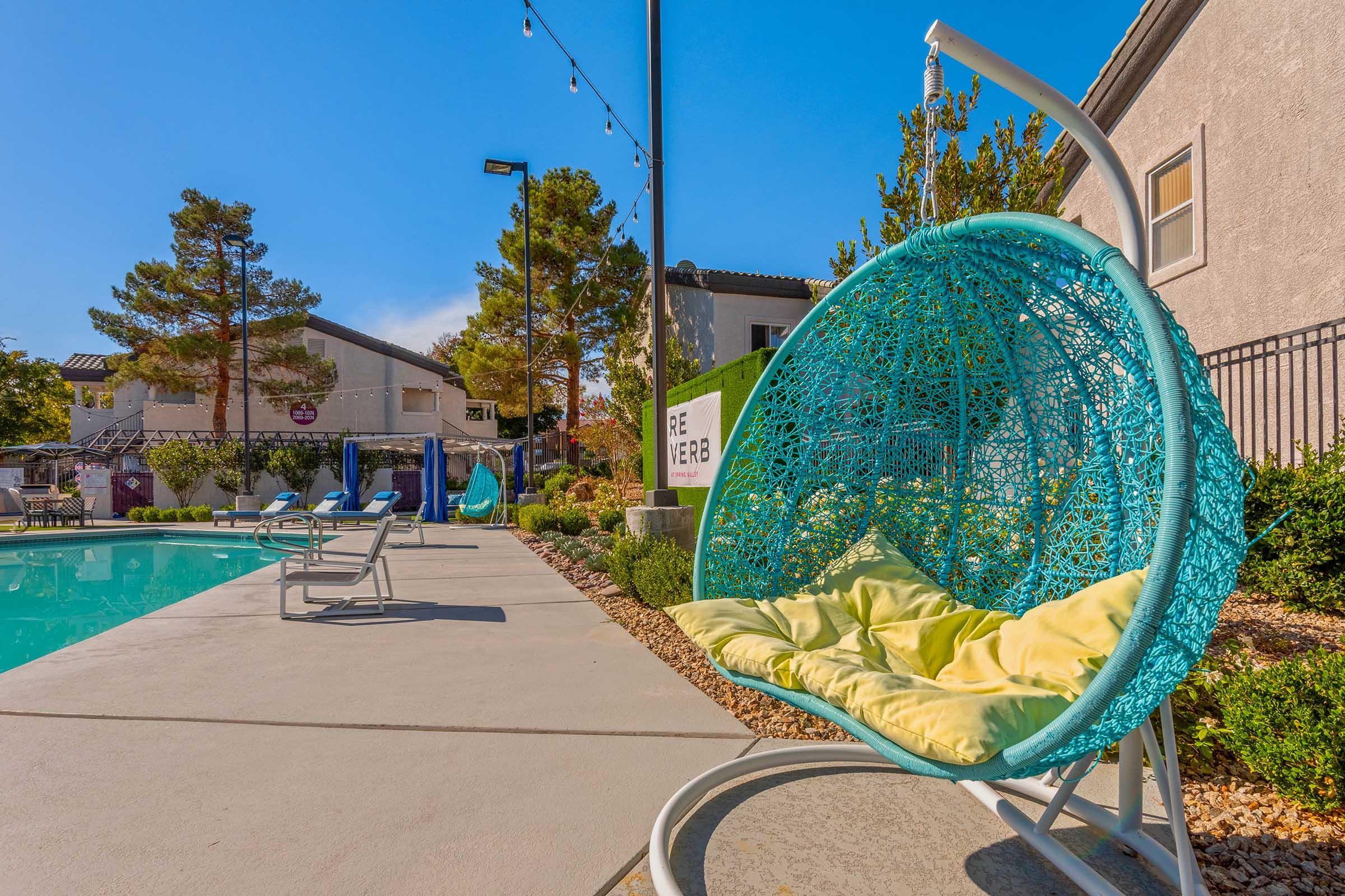 A turquoise hanging chair with a yellow cushion sits near a swimming pool. In the background, there are several lounge chairs and a building with greenery. The setting features clear blue skies and modern landscaping, creating a relaxing outdoor atmosphere.