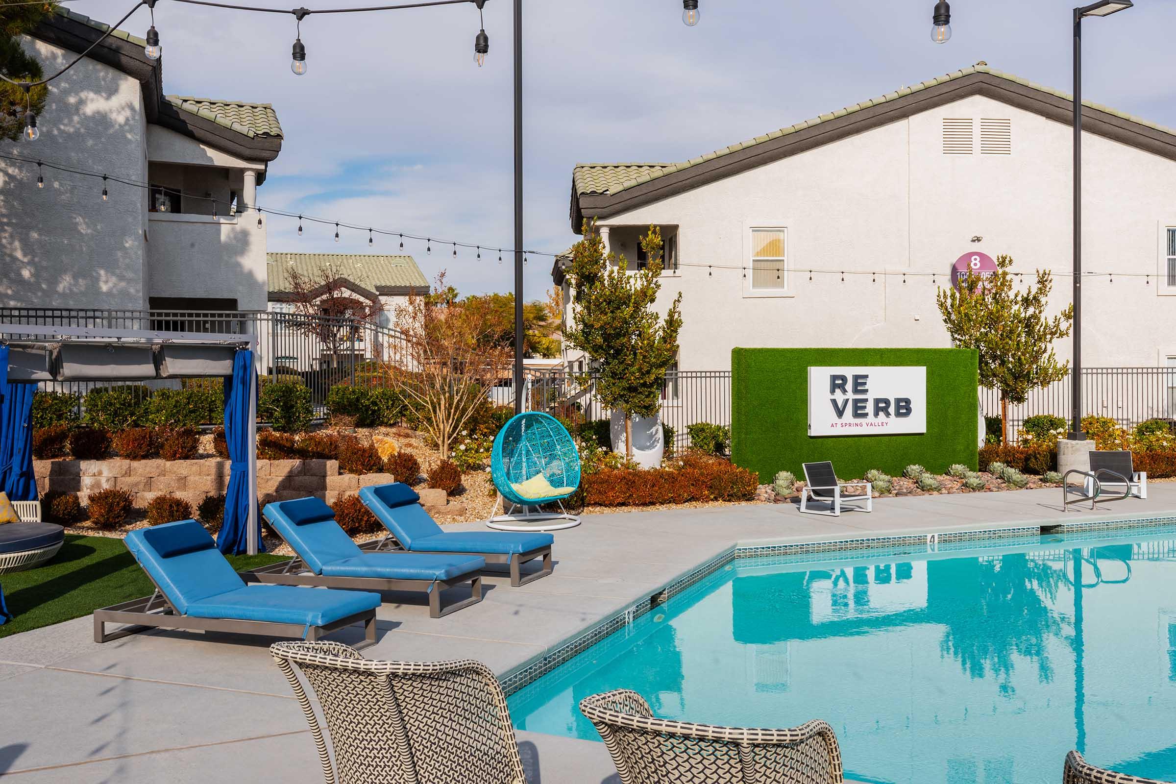 A modern outdoor pool area featuring lounge chairs, a vibrant green wall with the text "REVERB," and landscaped surroundings. There are decorative string lights overhead, and a cozy seating arrangement nearby. The atmosphere is inviting and perfect for relaxation.