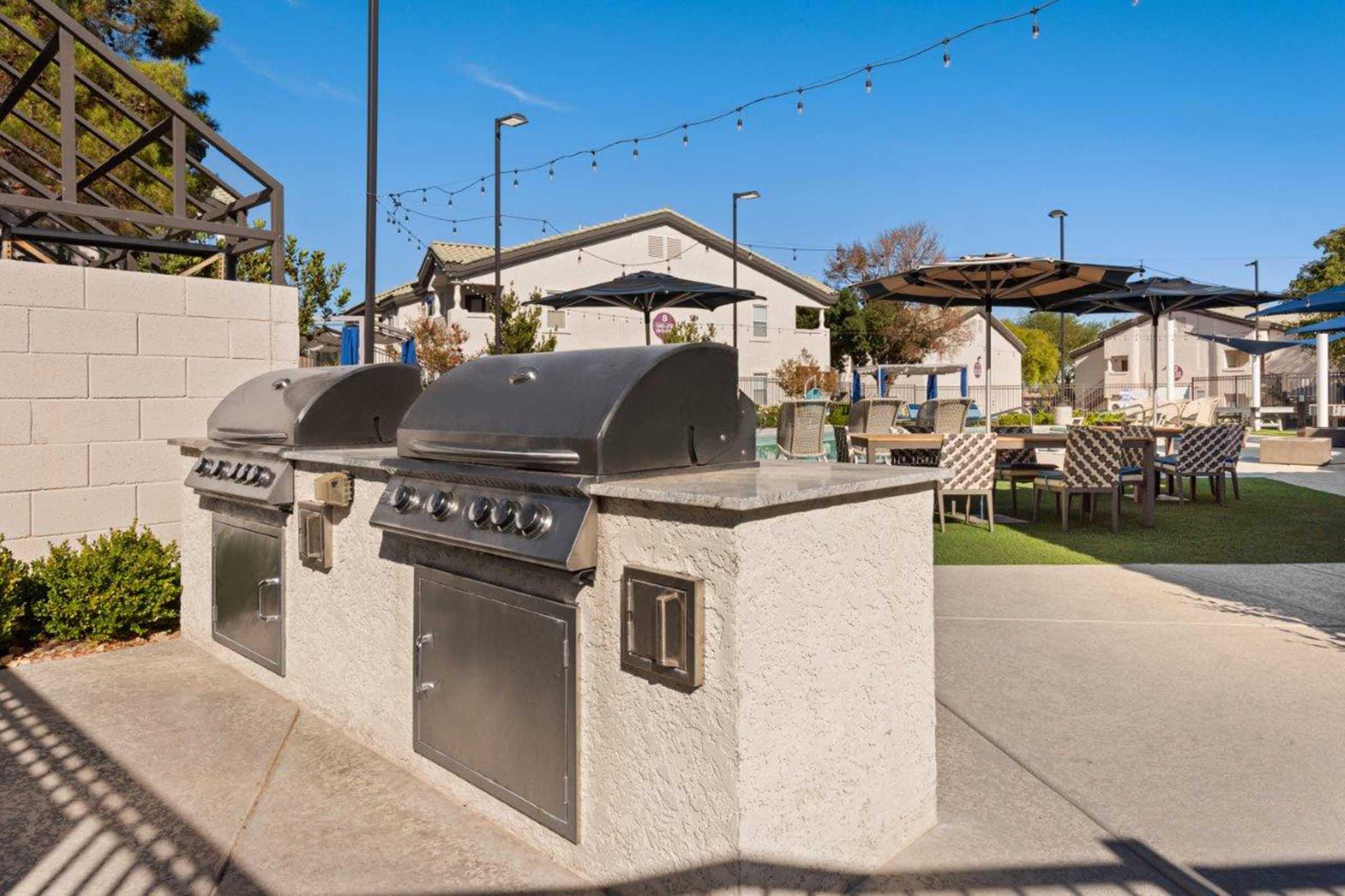 Two outdoor grills on a stone countertop in a landscaped area, surrounded by seating and table arrangements. A blue sky with a few clouds and string lights overhead enhance the inviting atmosphere of this recreational space.