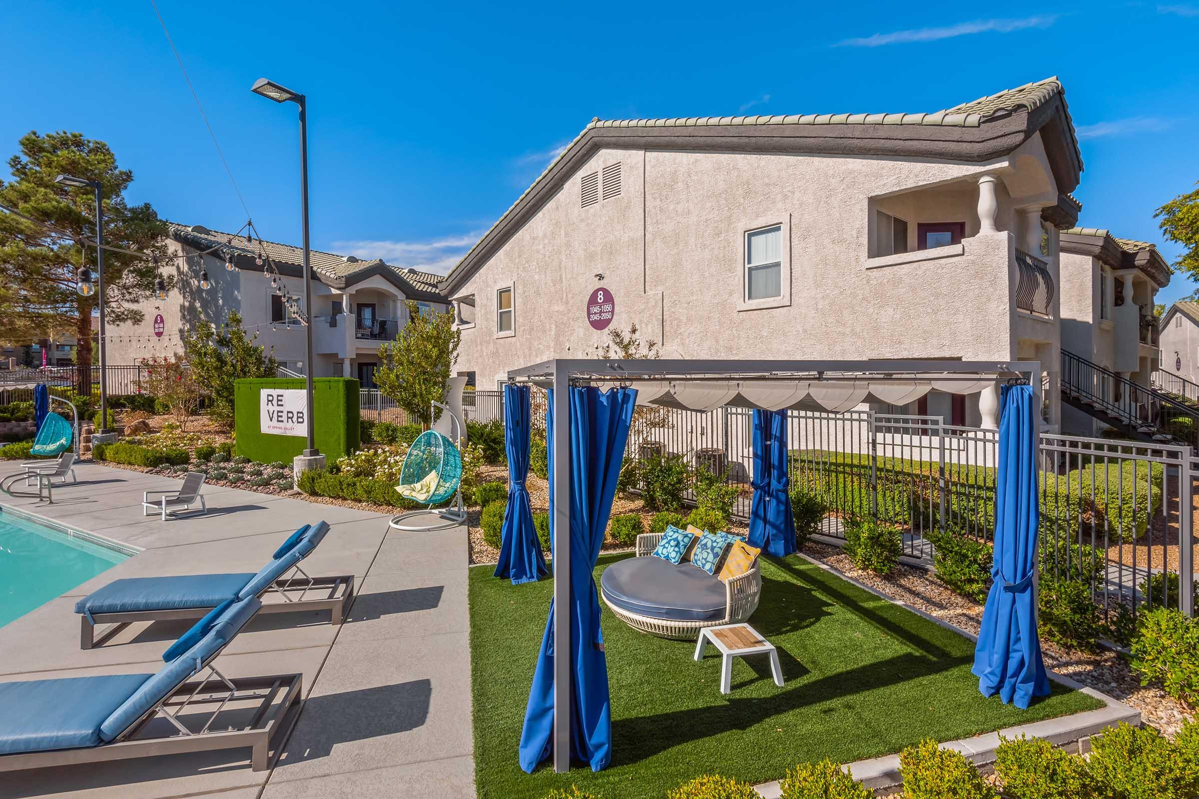A serene outdoor space featuring a swimming pool surrounded by lounge chairs, a shaded cabana with circular seating, and vibrant blue curtains. Lush greenery and well-kept landscaping enhance the inviting atmosphere, with modern apartment buildings in the background under a clear blue sky.