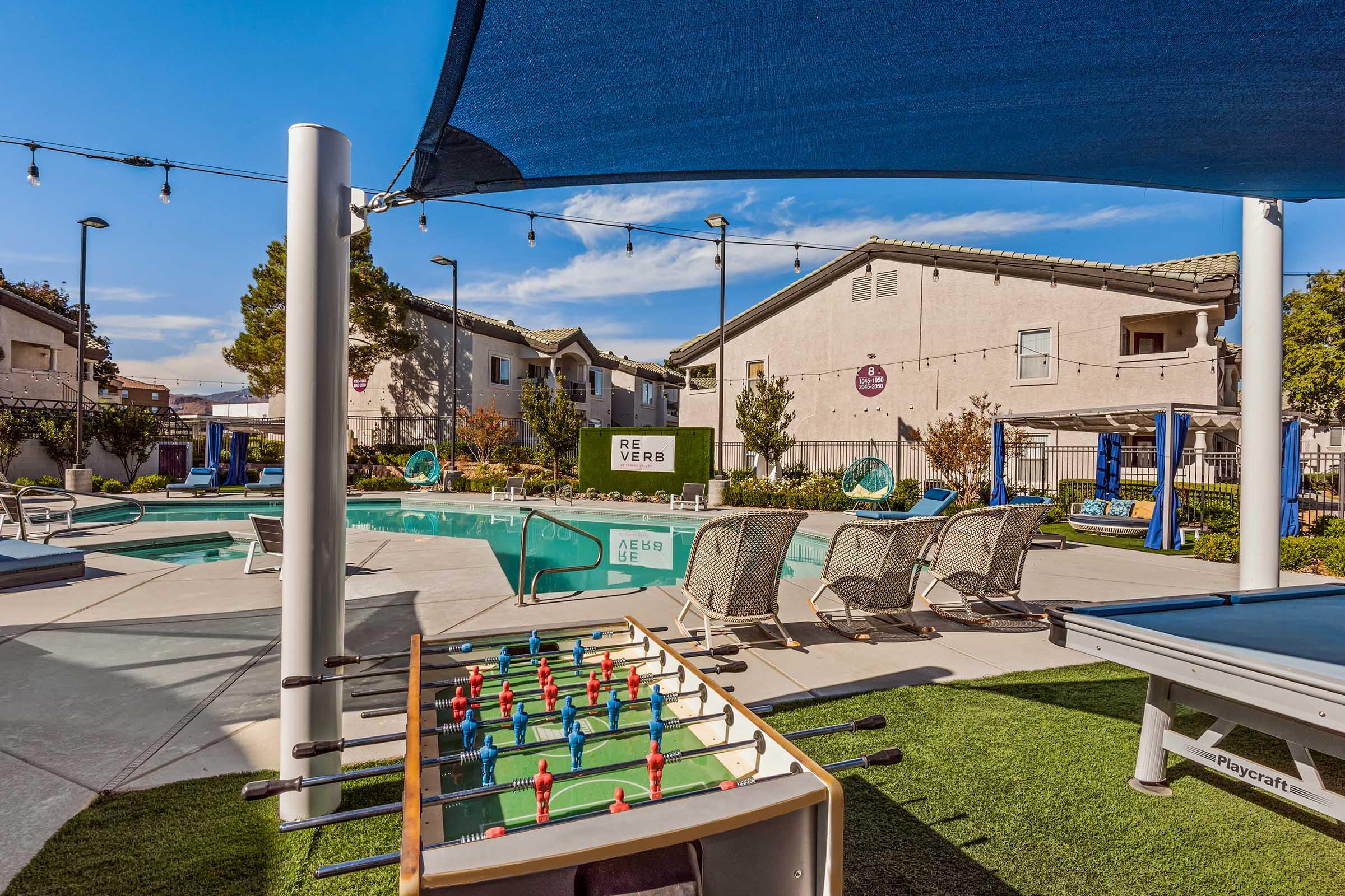 A vibrant outdoor leisure area featuring a swimming pool with lounge chairs, a shaded seating area, and games like foosball. Surrounding buildings add a residential feel, and string lights provide a warm ambiance. The scene is set for relaxation and entertainment in a sunny environment.