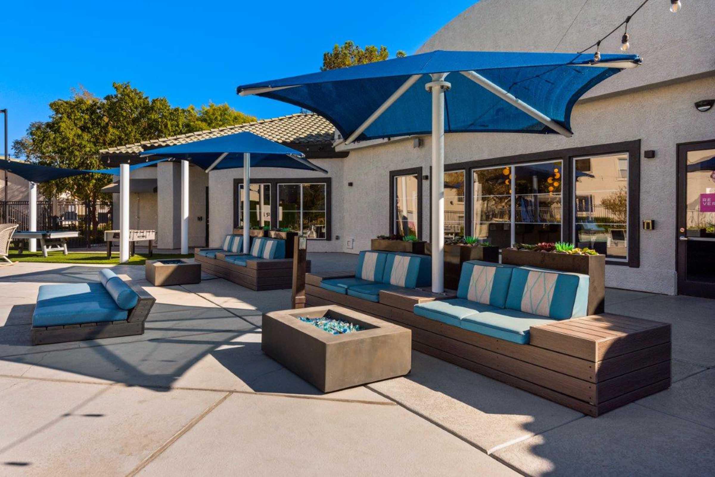 Outdoor lounge area featuring modern seating with blue cushions, large blue umbrellas providing shade, and a fire pit with a decorative center. The setting is bright with clear blue skies and a well-maintained environment, ideal for relaxation and social gatherings.