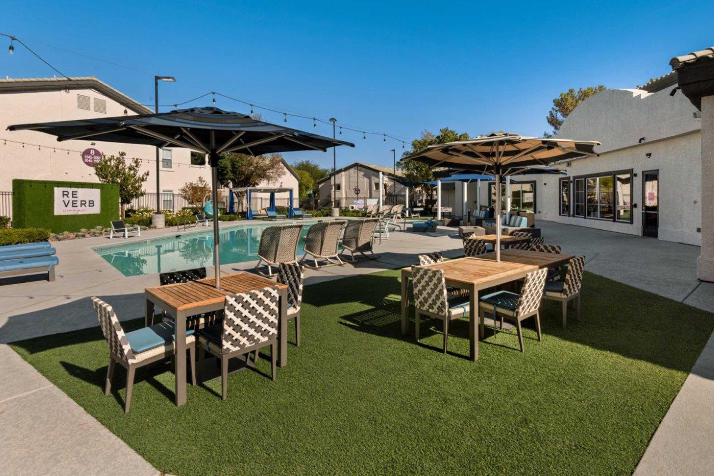 A modern outdoor pool area featuring lounge chairs, tables with umbrellas, and a well-maintained lawn. Surrounding the pool are cozy seating arrangements and a building in the background, creating a relaxing ambiance. The sky is clear, enhancing the inviting atmosphere.