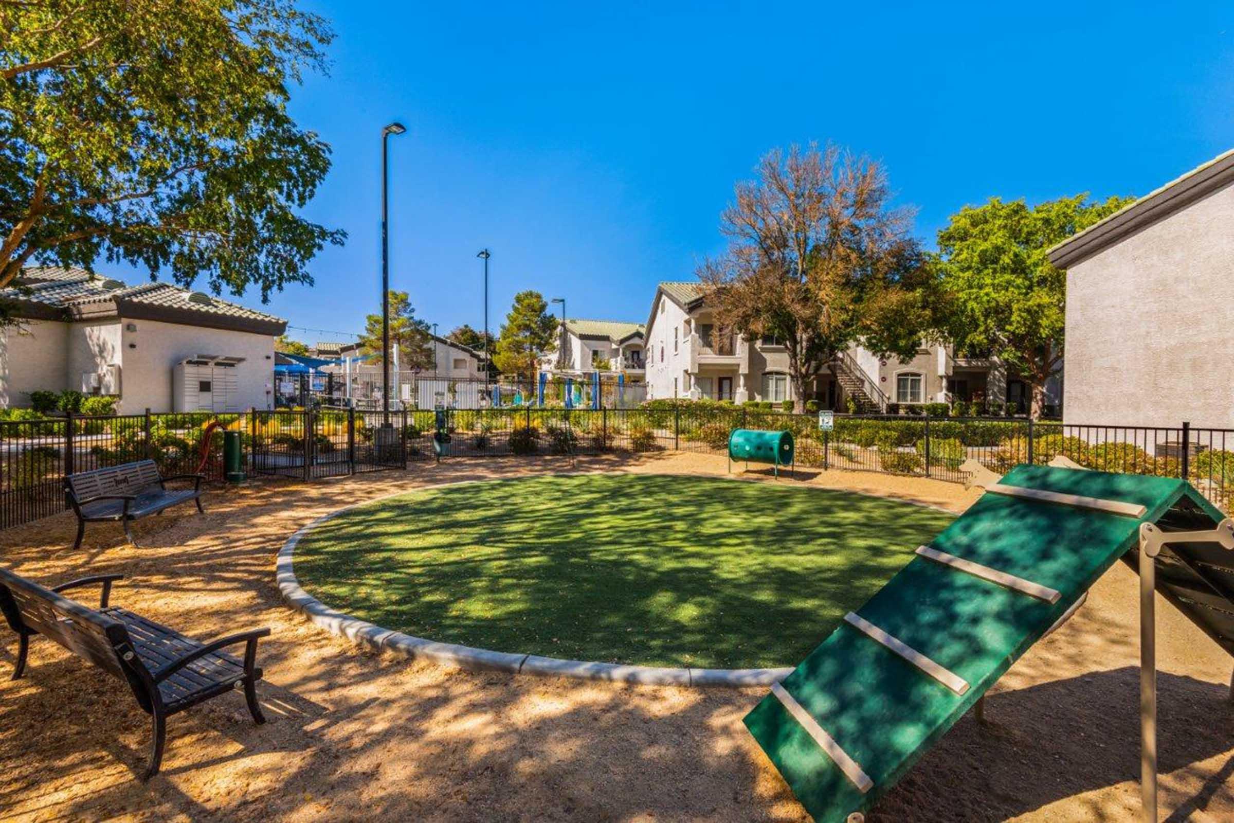A small park area featuring a circular patch of green grass, benches, and a slope for climbing. In the background are residential buildings and trees under a clear blue sky. The park appears well-maintained and inviting for leisure activities.