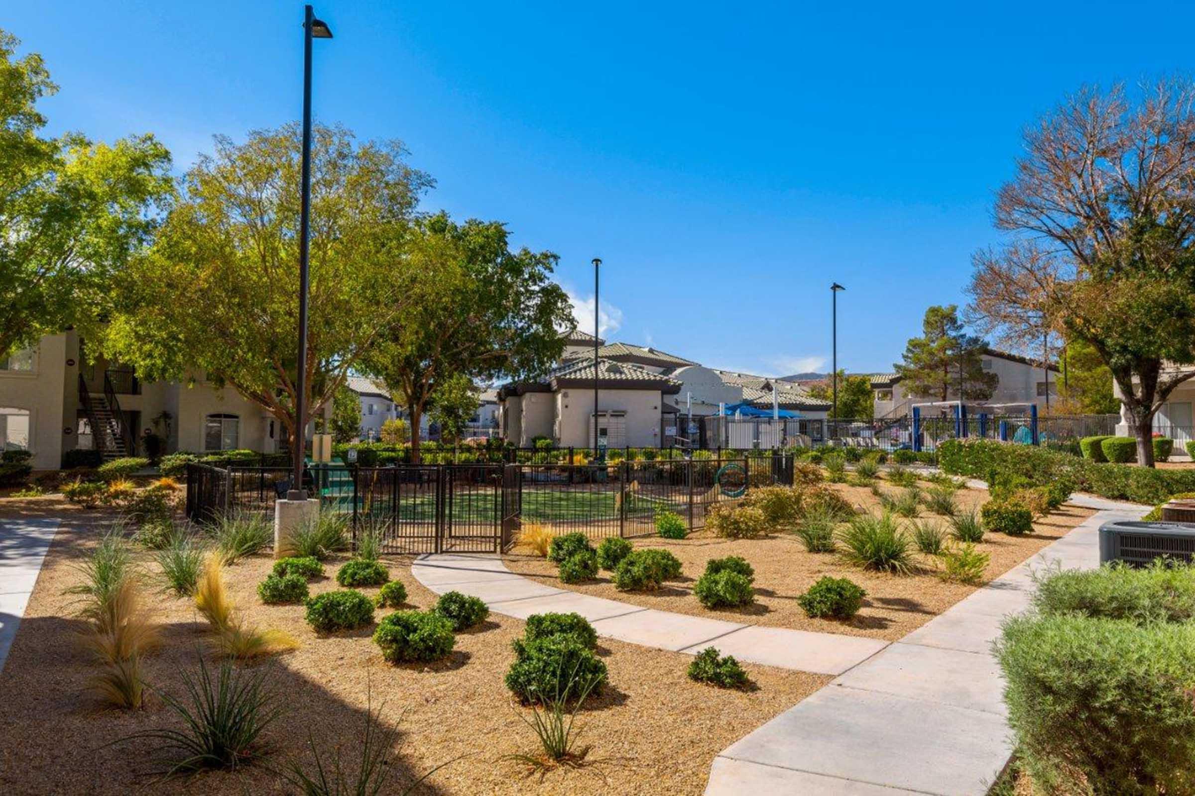 A landscaped park area featuring a fenced playground, surrounded by bushes and trees. The sky is clear and blue, with several lamp posts visible. In the background, there are residential buildings and some recreational facilities. The ground is covered with gravel and shrubs, creating a peaceful outdoor space.