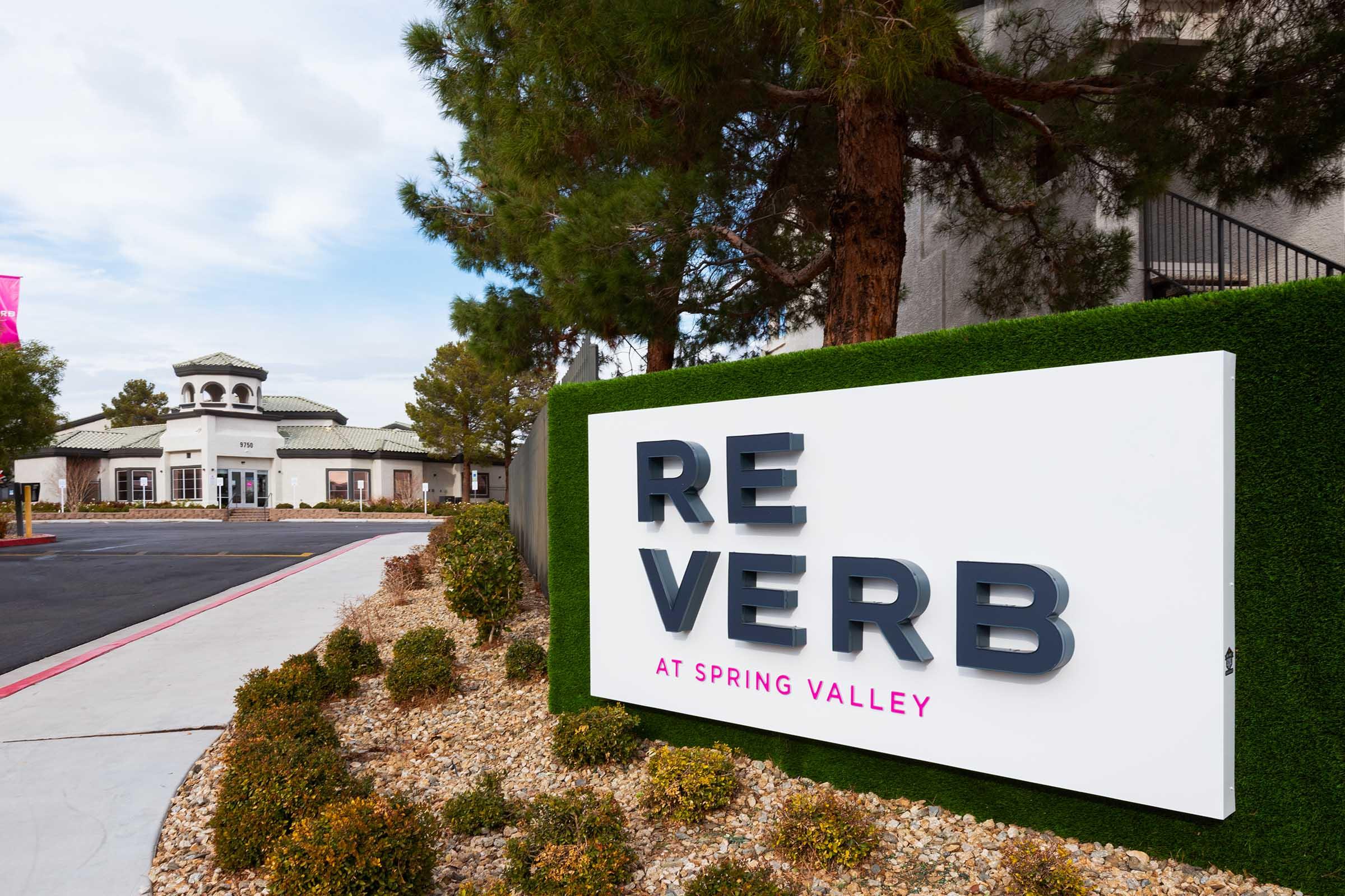 Sign for "REVERB at Spring Valley" positioned near the entrance, surrounded by landscaped greenery. A building is visible in the background, with a cloudy sky overhead. The area features neatly arranged shrubs and a paved path leading towards the structure.
