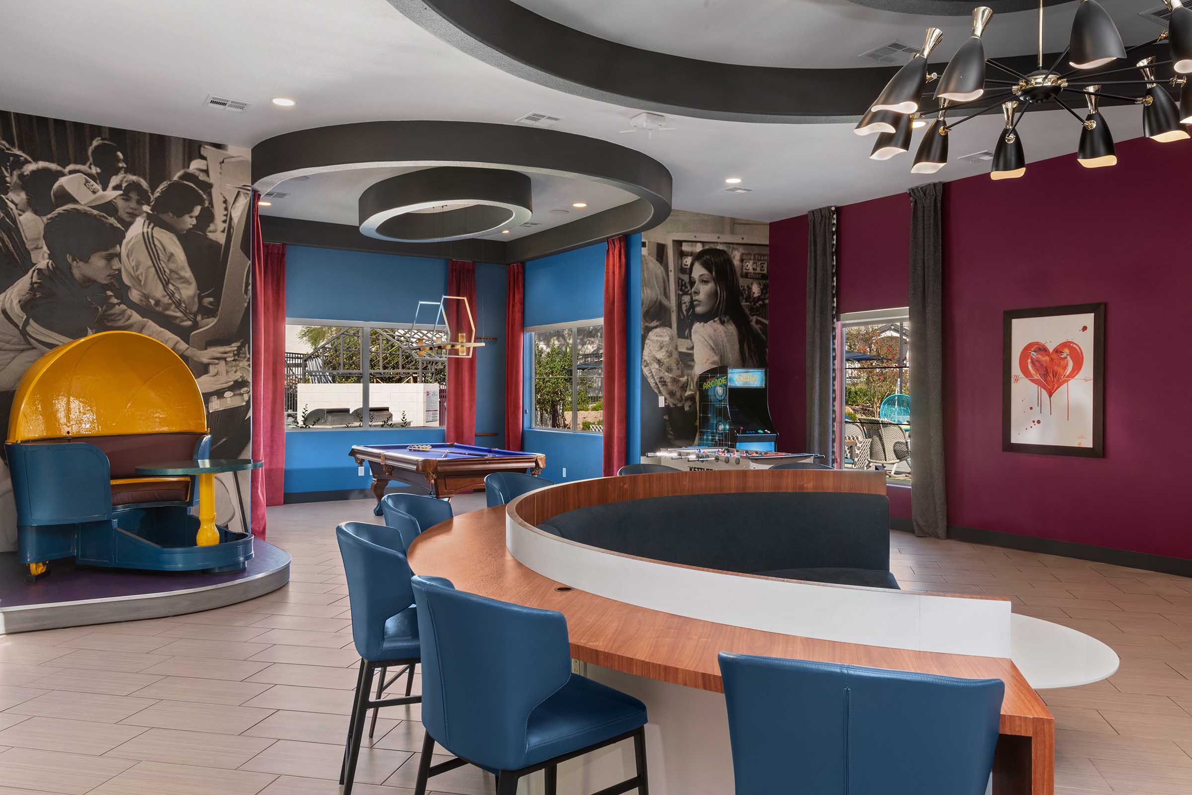 Modern lounge area featuring a curved counter and blue chairs, with a vibrant color palette including maroon and blue walls. The decor includes a large mural of people, artwork on the walls, and a decorative light fixture. A pool table is visible in the background, creating a relaxed social atmosphere.