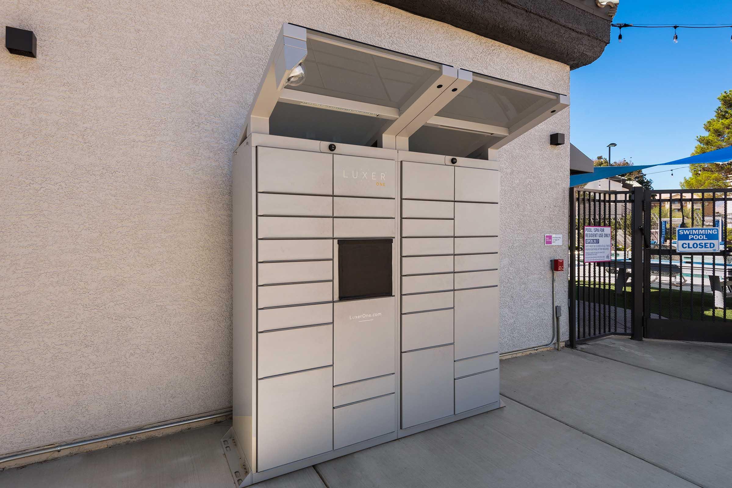 A modern automated package delivery locker system with a sleek design, situated against a textured wall. The lockers feature multiple compartments for secure storage of parcels. A sign indicates that the area is monitored, and there are access control measures in place, such as a gated entrance nearby.