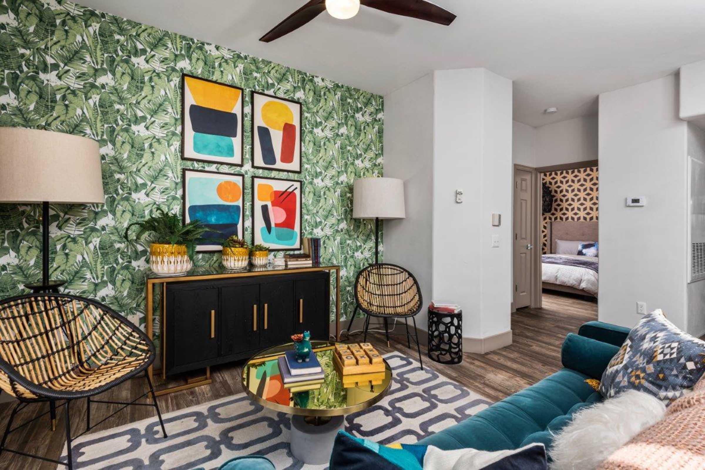 A stylish living room featuring a bold green botanical wallpaper, colorful abstract art on the wall, a dark wood sideboard with decorative items, two round lamps, a round glass coffee table with books, and modern seating arrangements including a teal sofa and woven chairs.