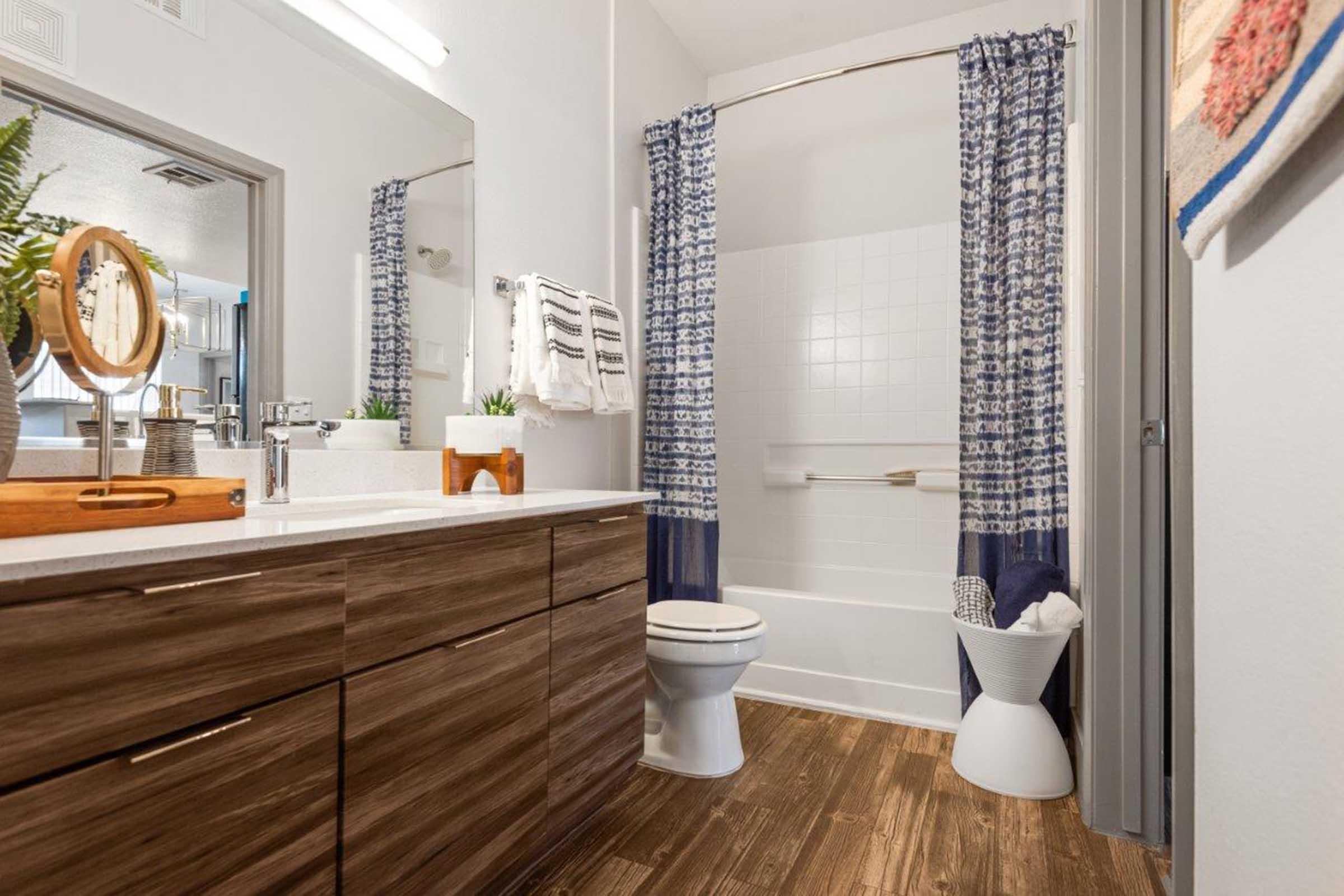 A modern bathroom featuring a wooden vanity with a sink, large mirror, and decorative plants. The bathtub is enclosed with blue patterned shower curtains. A towel rack is mounted on the wall, and a white stool is placed beside the tub, contributing to a clean and contemporary aesthetic.