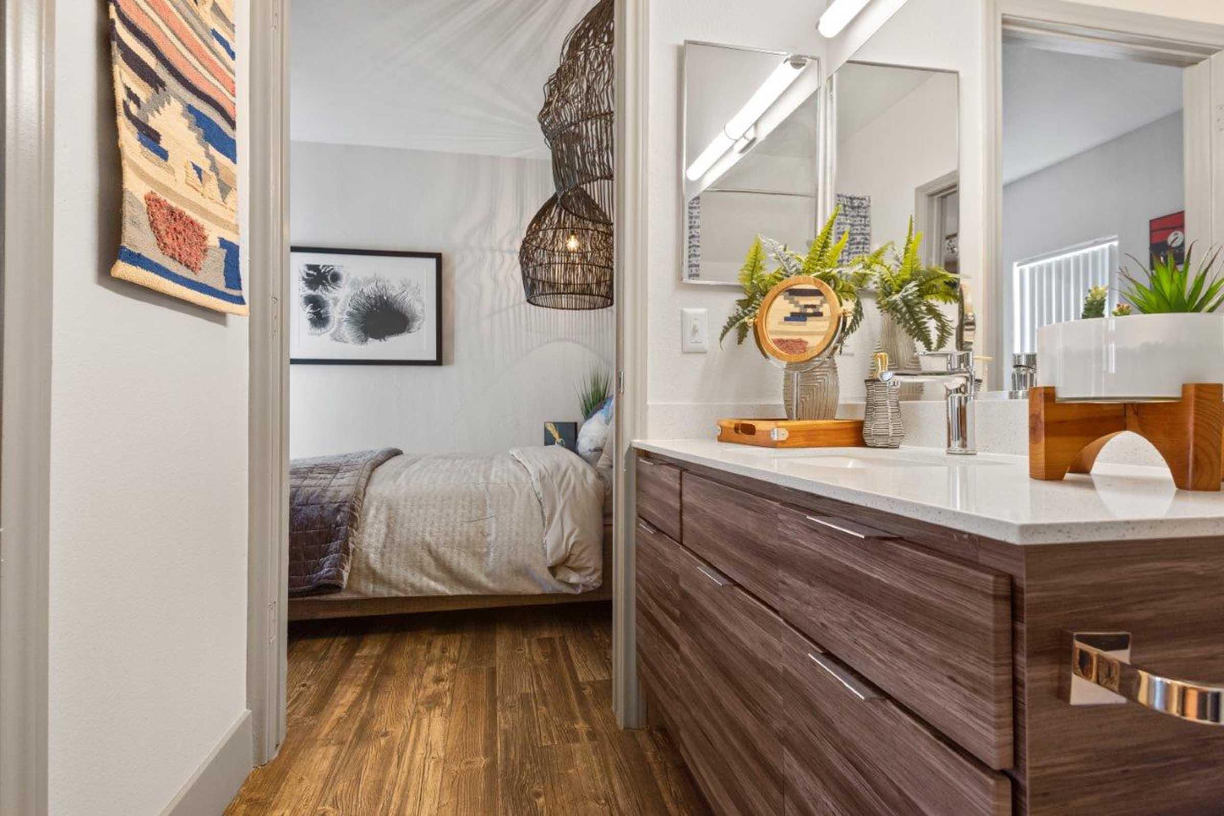 Modern bathroom with a wooden vanity and a sink, featuring a round mirror above. Warm-toned wooden flooring. In the background, a softly lit bedroom with a bed and framed artwork. Stylish hanging light fixtures add ambiance, while decorative plants and accessories enhance the contemporary decor.