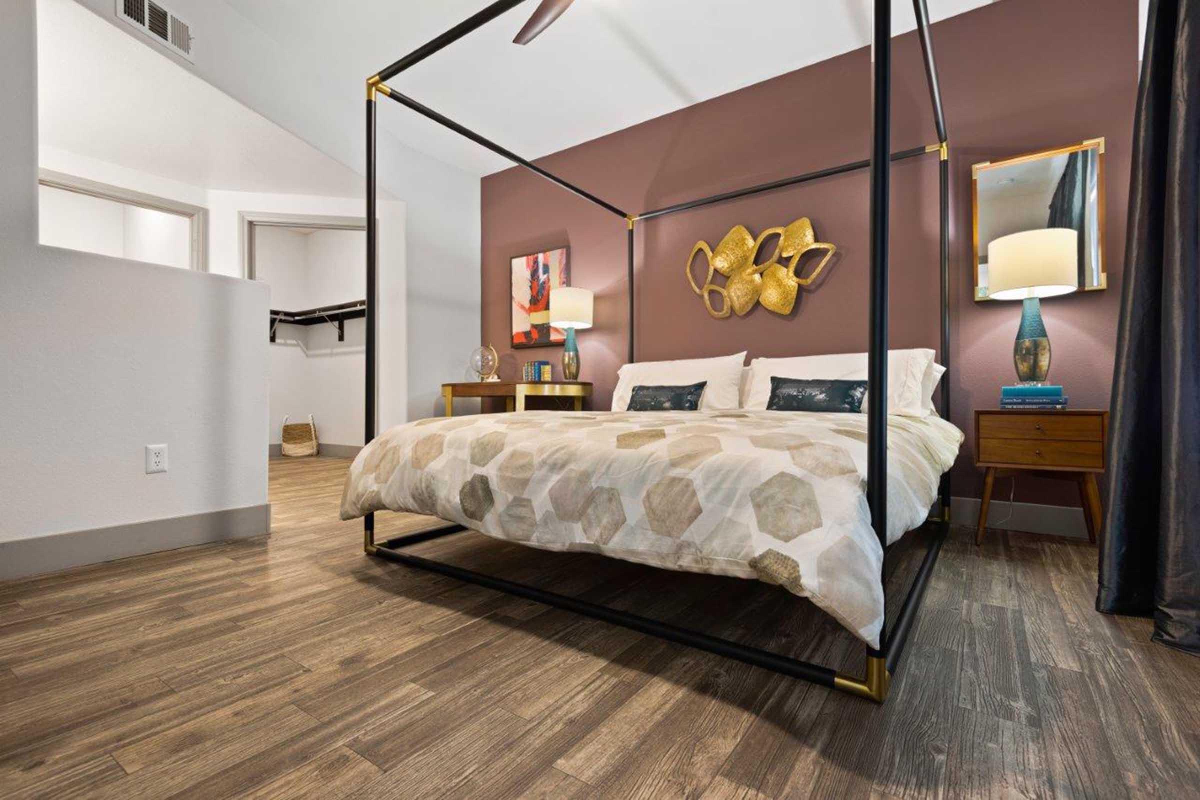 A modern bedroom featuring a four-poster bed with a geometric-patterned blanket. The walls are painted a warm brown hue, complemented by decorative wall art. A side table holds a lamp and a plant, while wooden flooring adds warmth. A closet is visible in the background, enhancing the room's organized look.