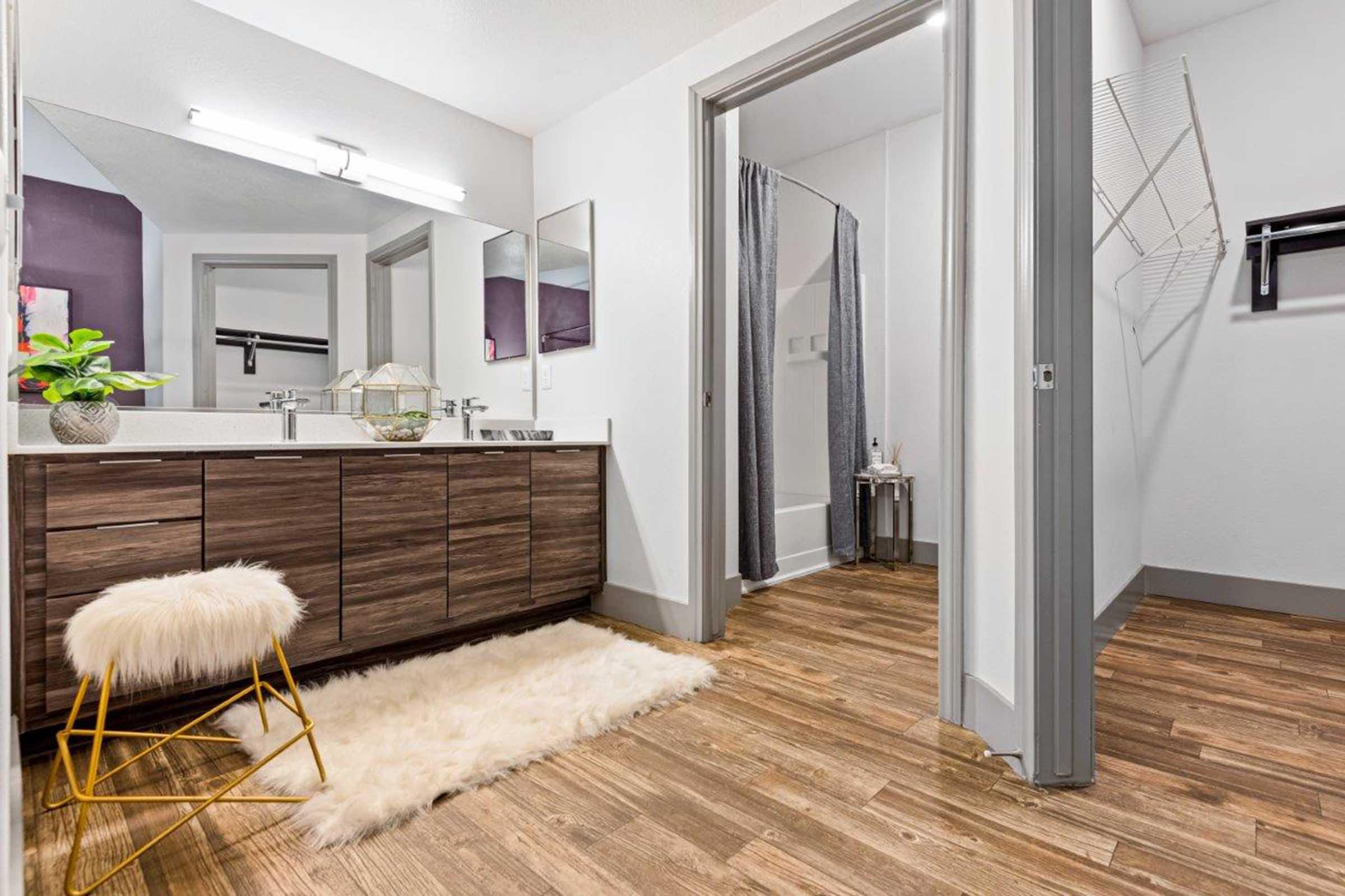 Modern bathroom featuring a wooden vanity with a double sink, a plush white rug, and a stylish gold stool. Mirrors and soft lighting enhance the space, while a shower area with a curtain and a clothing rack are visible, creating a clean and inviting atmosphere.