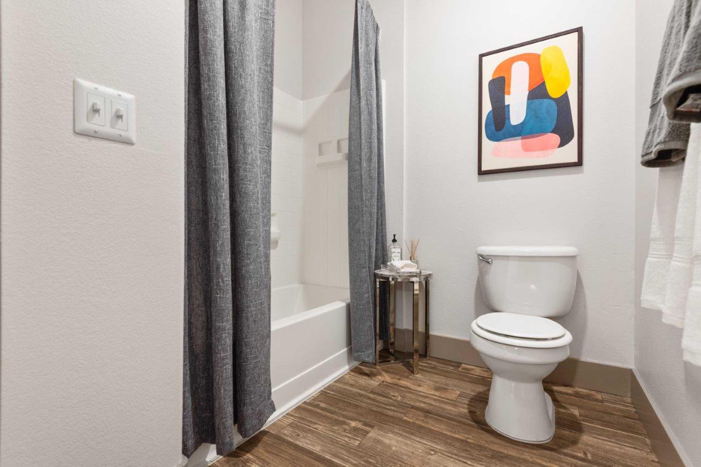 A modern bathroom featuring a white toilet, a bathtub with a curtain, and a small side table. The walls are light-colored, and a colorful abstract artwork hangs above the table. Soft towels are neatly arranged on a towel rack, and the flooring is a brown wood-like material.