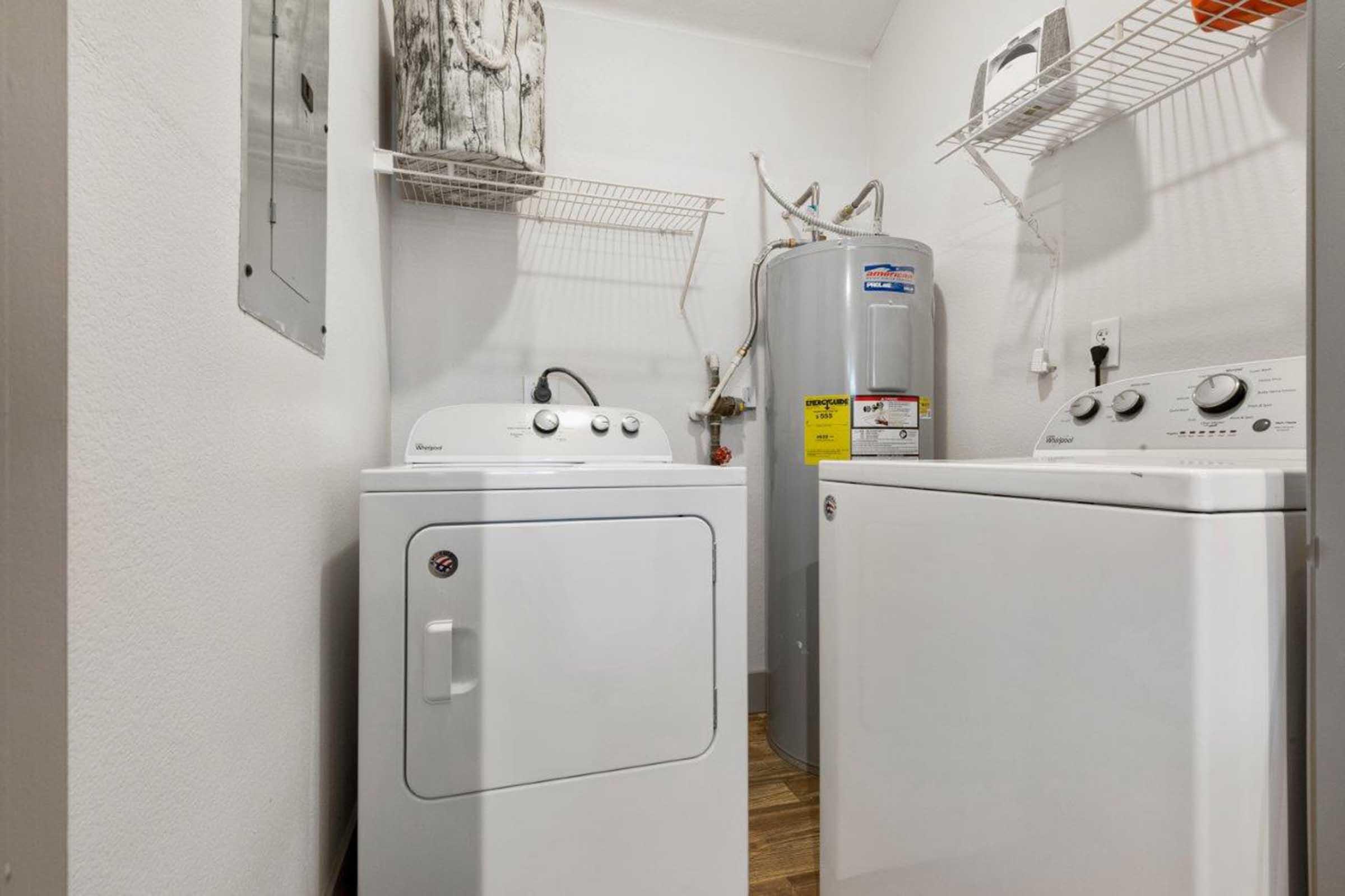 A laundry room featuring a white washer and dryer set side by side. A water heater is visible on the right, along with a wall shelf above the appliances. The room has light-colored walls and a wooden floor, creating a clean and organized space.