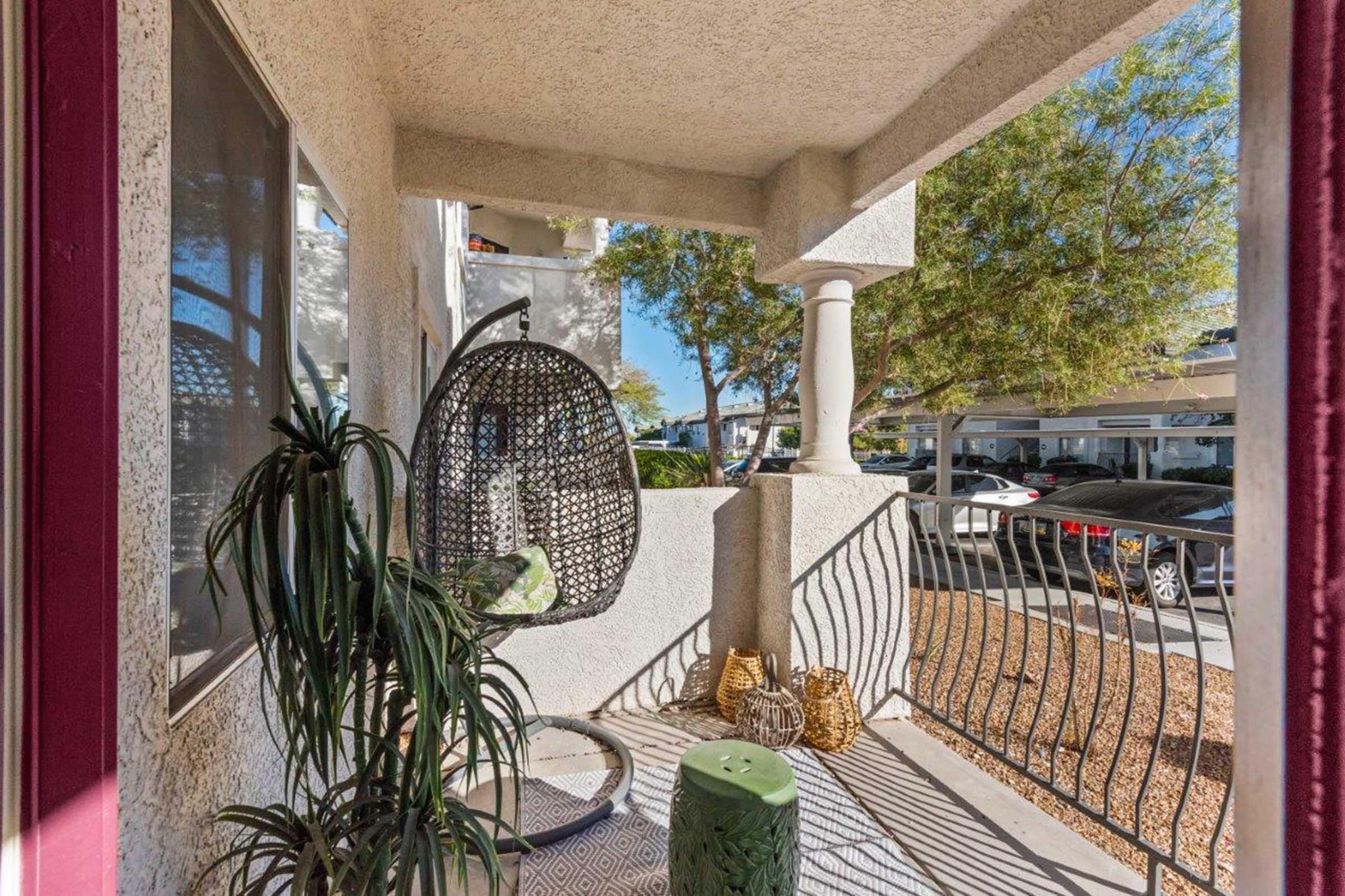 A cozy patio area featuring a hanging wicker chair, a green decorative stool, and potted plants. The space is surrounded by a railing and has a view of parked cars and trees outside. The entrance is characterized by a warm, inviting atmosphere with natural light.