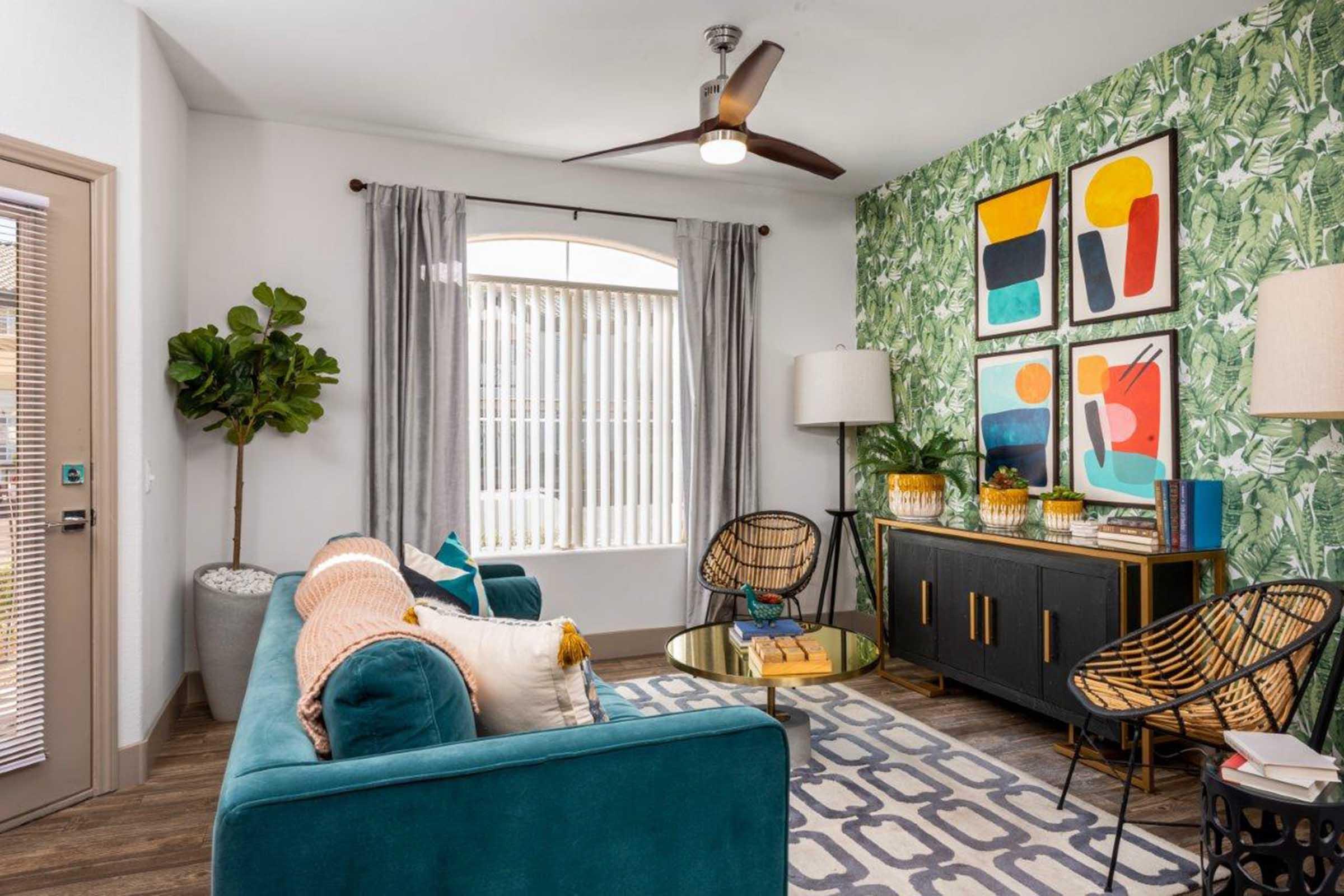Modern living room featuring a green and white leaf-patterned accent wall. There are two stylish chairs and a round coffee table in front of a plush teal sofa with decorative pillows. Bright artwork hangs on the wall. Natural light streams in through a large window adorned with vertical blinds.