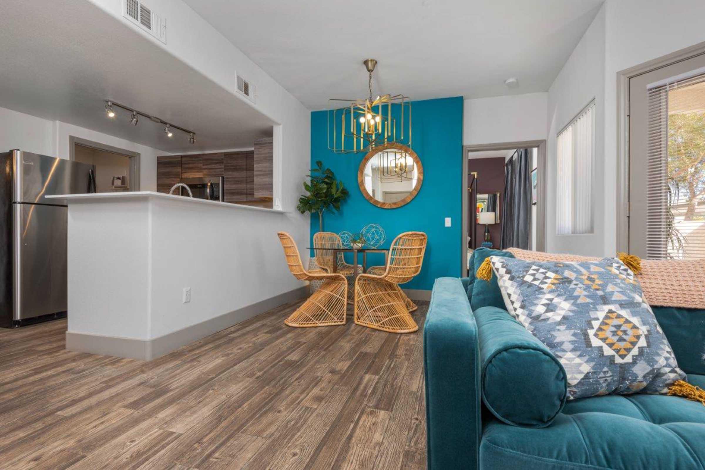 A modern living space featuring a turquoise accent wall, a dining area with a round table and rattan chairs, and a cozy couch adorned with decorative pillows. The kitchen area is visible with stainless steel appliances, while natural light flows through the windows, enhancing the welcoming atmosphere.