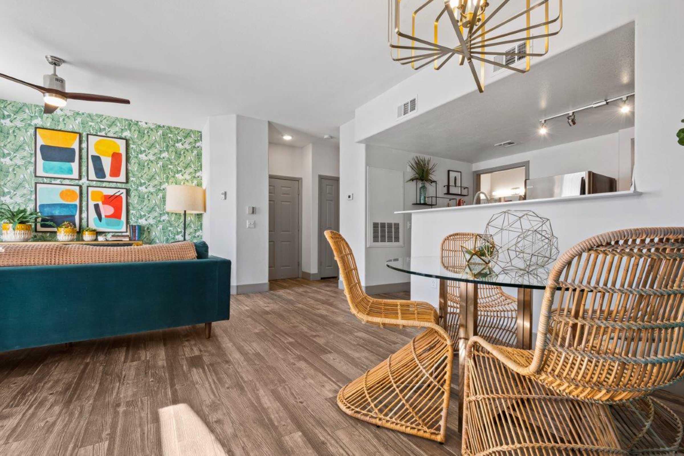 Modern living room featuring a teal sofa and a vibrant green patterned accent wall with framed art. A stylish dining area with a glass table and wicker chairs is visible. Natural light fills the space, highlighting the wood flooring and contemporary decor elements.