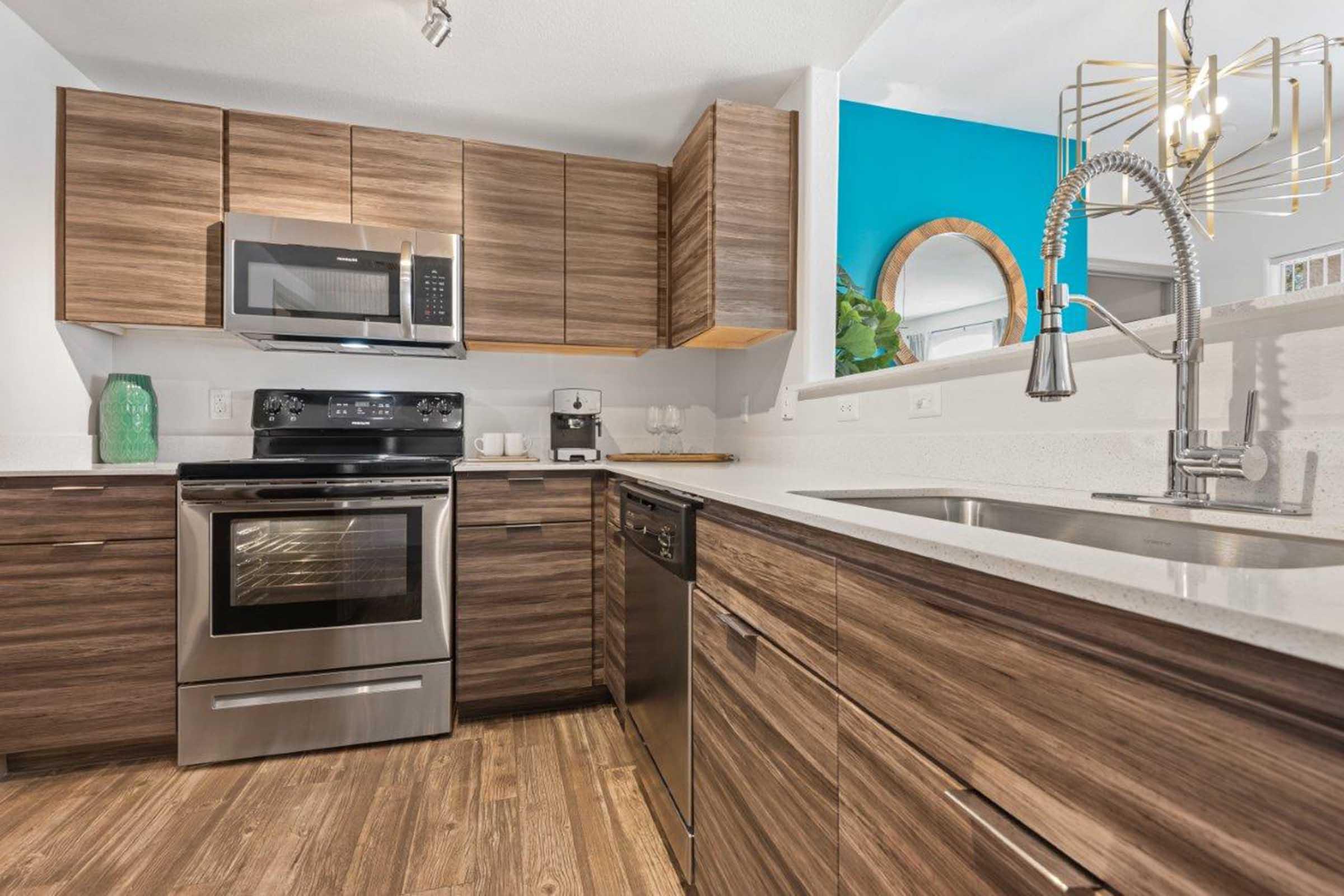 Modern kitchen featuring wooden cabinets, a stainless steel refrigerator and oven, a sink with a sleek faucet, and a light countertop. The walls are painted in a vibrant blue, and there's a circular mirror and green decorative items adding a touch of style. Well-lit and designed for functionality.