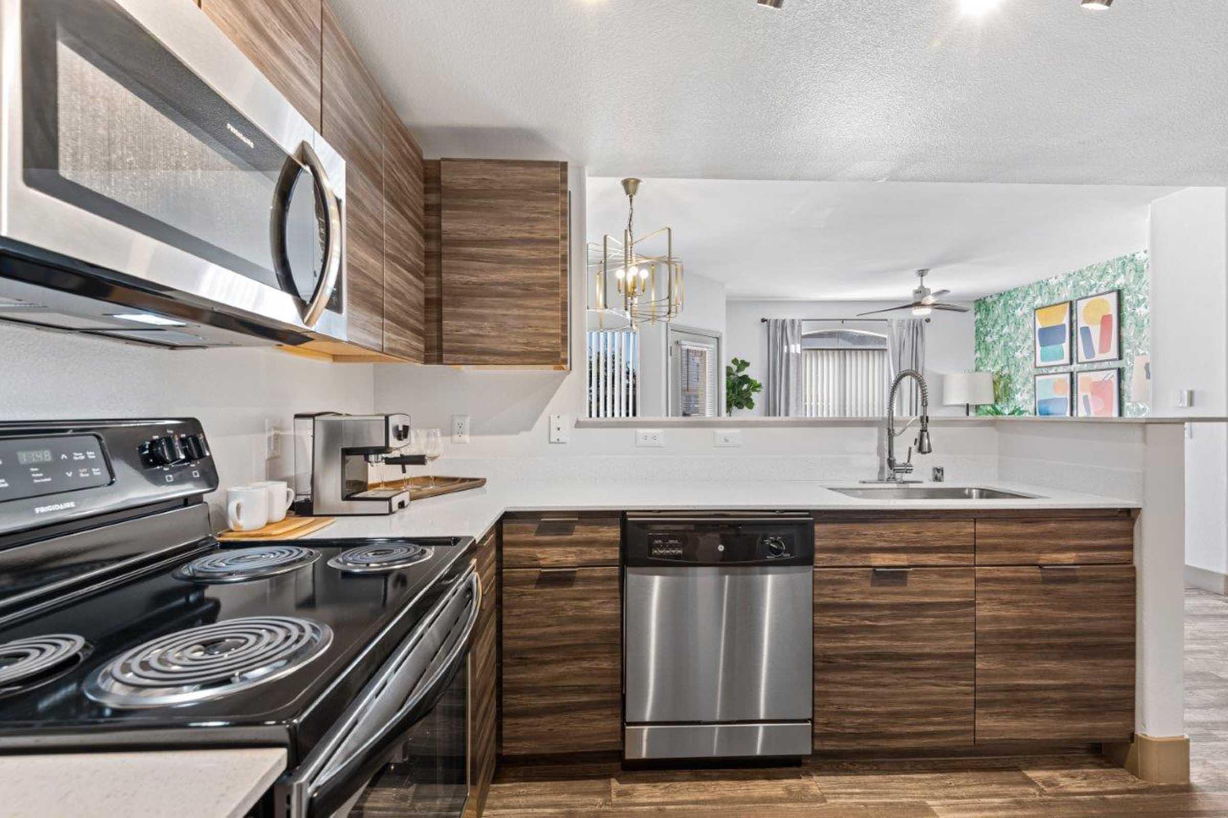Modern kitchen featuring wooden cabinetry, stainless steel appliances including a microwave, stove, and dishwasher. The countertop is equipped with a sink, and there is a light fixture above. The background shows a living area with greenery and colorful artwork. Natural light enters through a window.