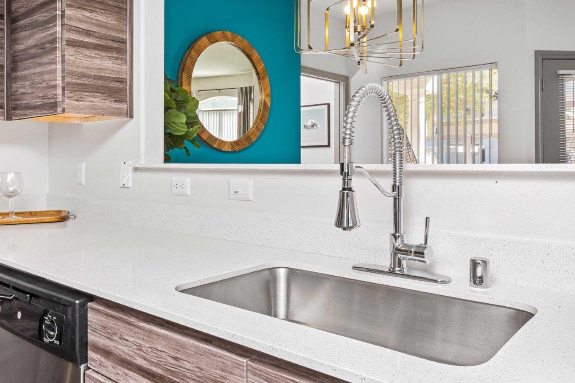 Modern kitchen featuring a sleek stainless steel sink with a chrome faucet, white countertop, and dark wood cabinetry. A round mirror with a wooden frame is mounted on the wall, alongside a vibrant turquoise accent wall. A decorative plant and stylish light fixture complete the contemporary design.