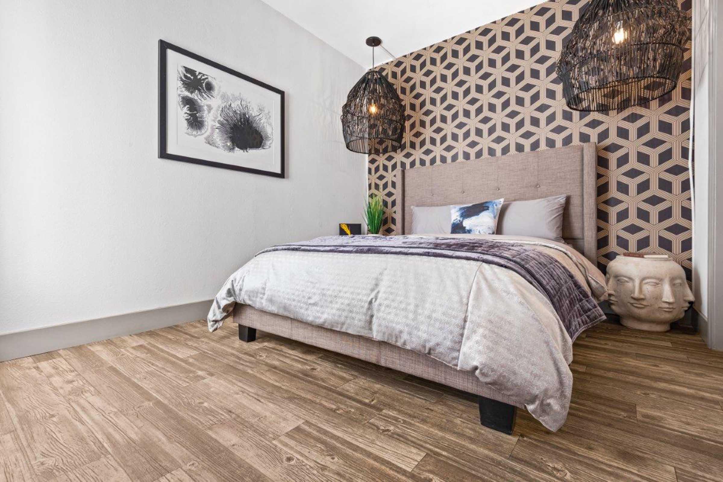 A modern bedroom featuring a bed with gray bedding, geometric patterned wallpaper, and two stylish pendant lights. A decorative headboard is behind the bed, with a framed artwork on the wall. The floor has wooden planks, and a decorative sculpture is placed beside the bed. A touch of greenery adds freshness to the space.
