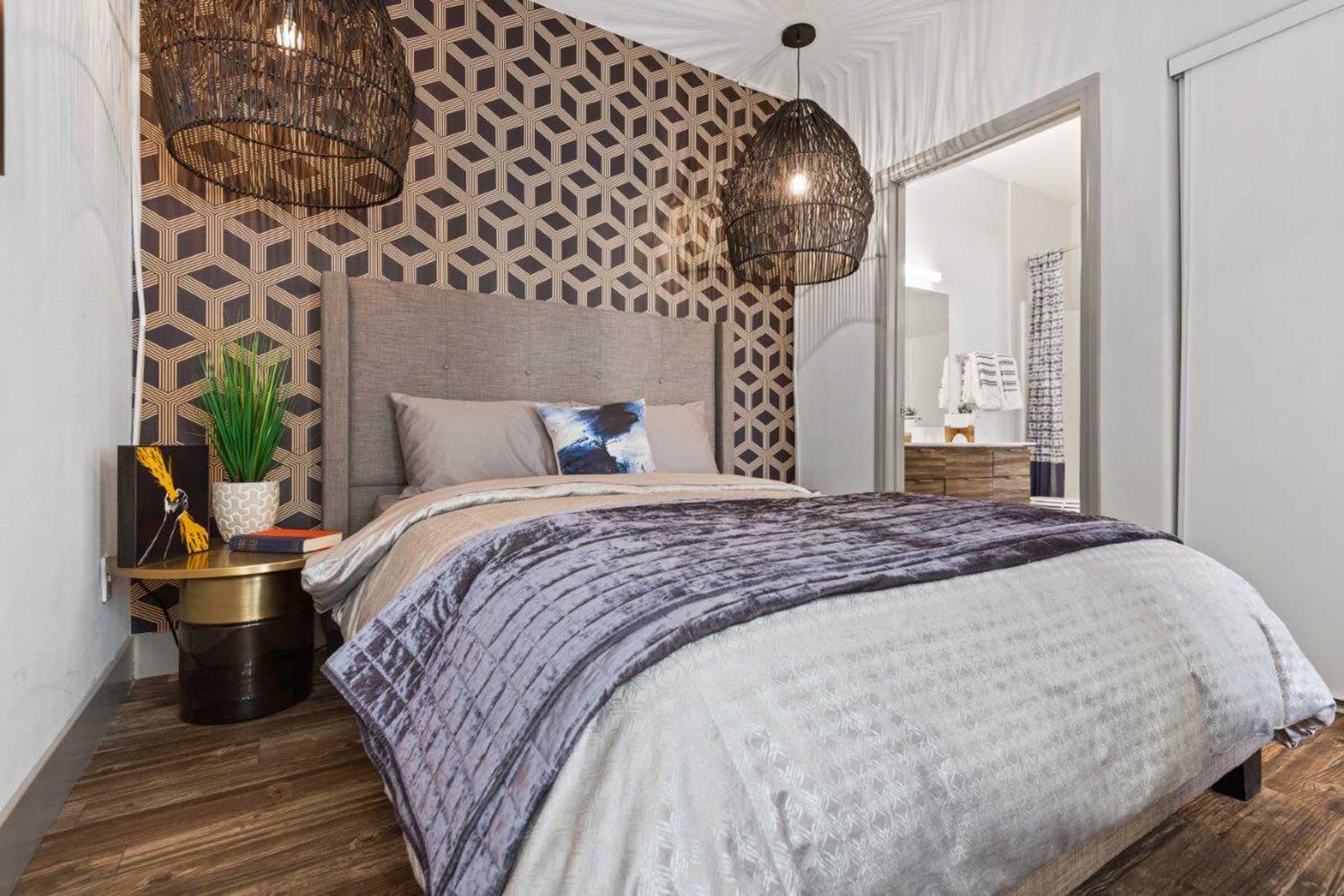 A stylish bedroom featuring a geometric patterned accent wall in muted tones. The room has a cozy bed with soft bedding, a decorative pillow, and a bedside table with a plant and a book. Two large, modern pendant lights hang above, creating a warm atmosphere, while a bathroom door is visible in the background.